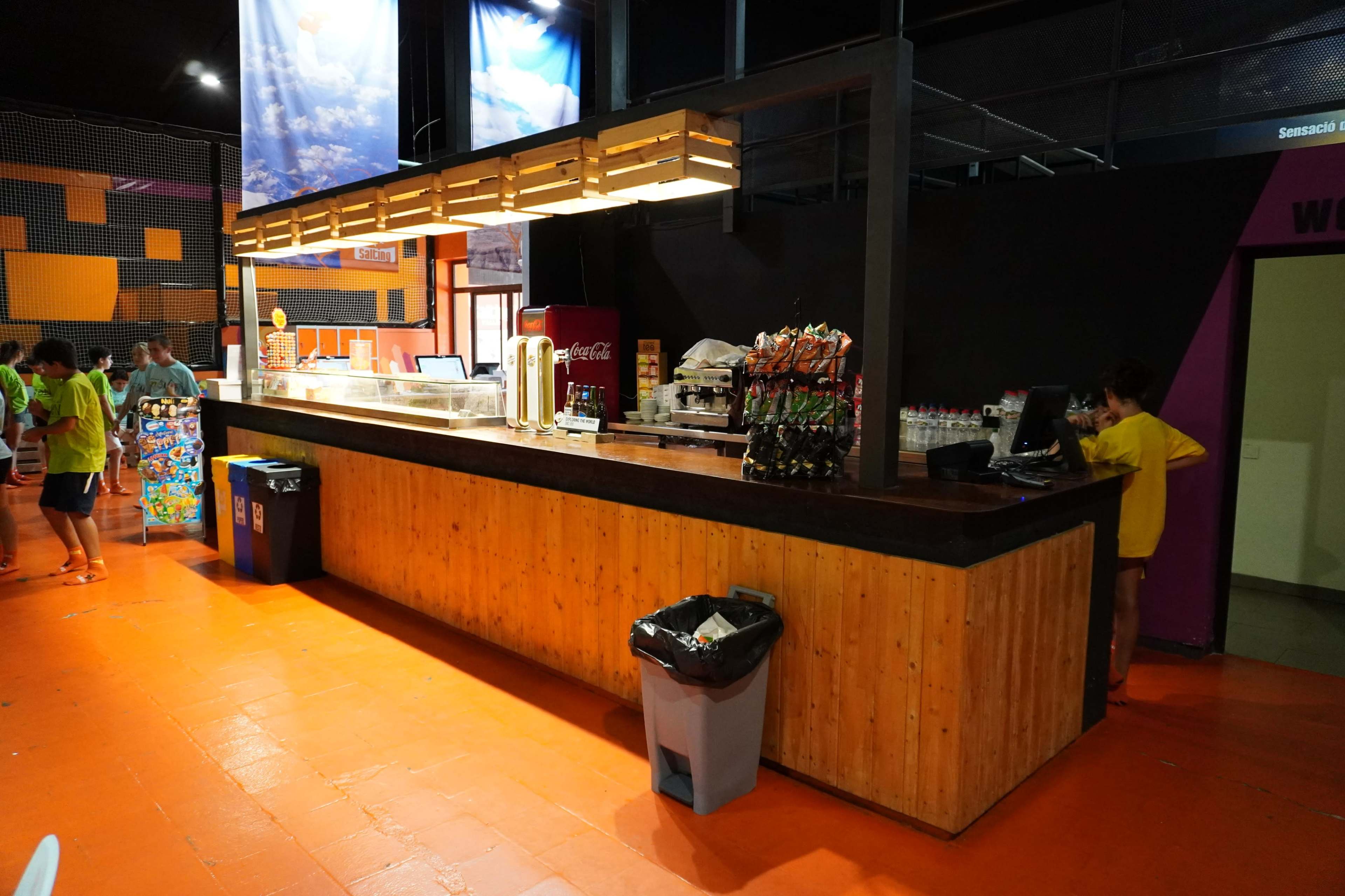 A brightly lit concession area with a wooden counter and various snack and drink options displayed, along with two trash bins nearby.