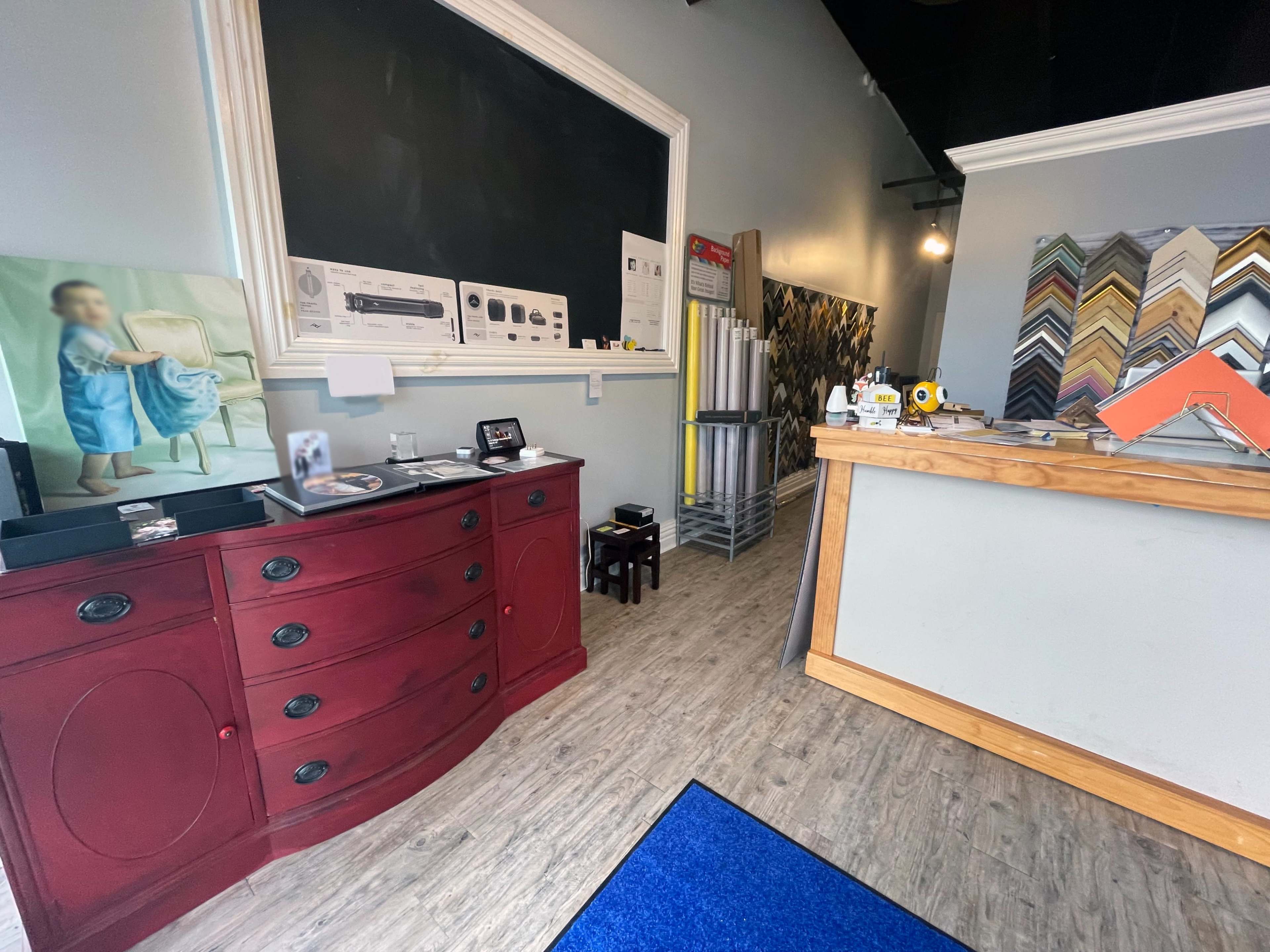 The image shows the interior of a frame shop featuring a red display cabinet, a service counter, and various framing supplies on a wall.