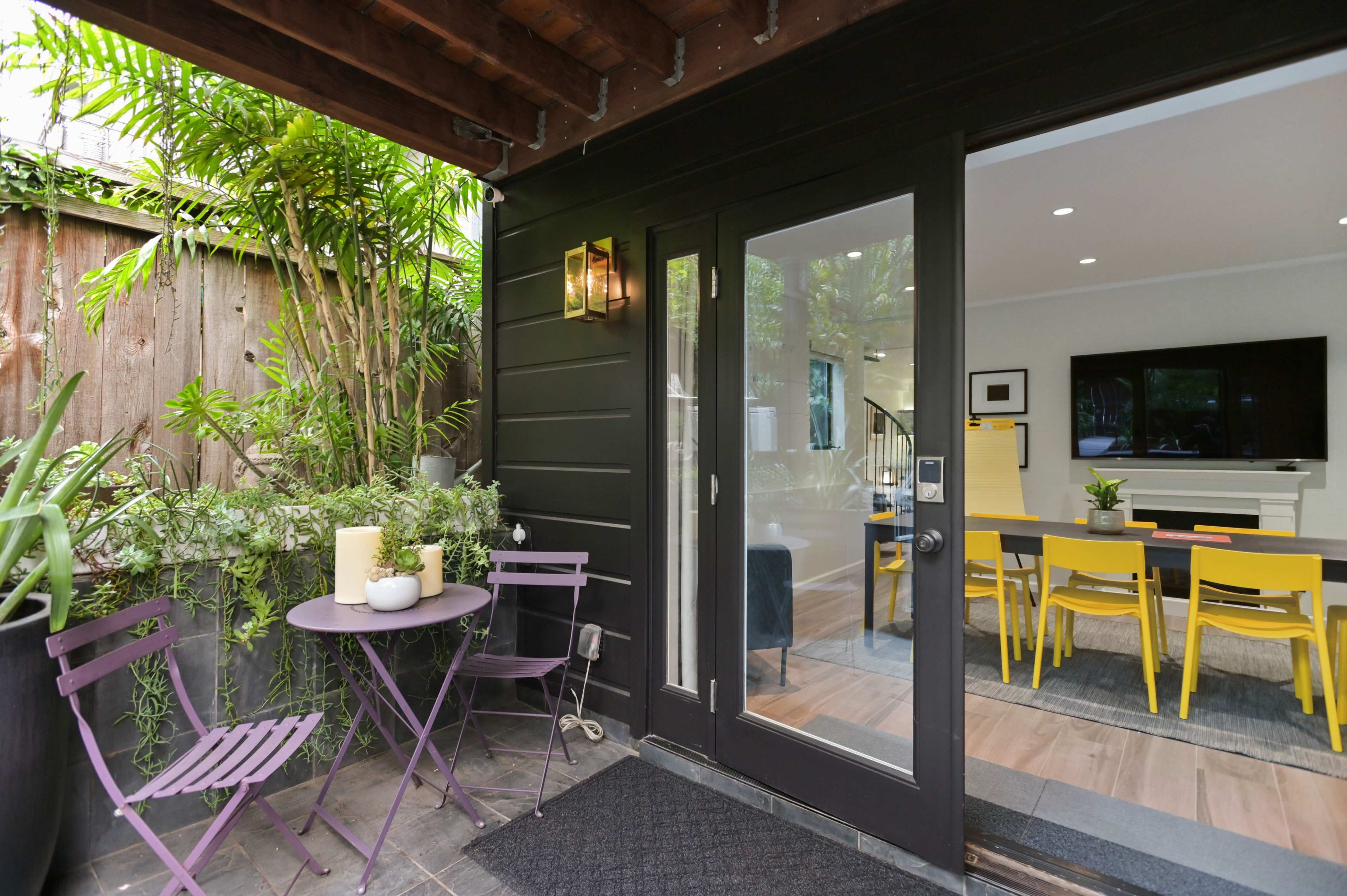 A small outdoor seating area with purple metal chairs and a table is adjacent to a modern dining space featuring a large TV and yellow dining chairs.