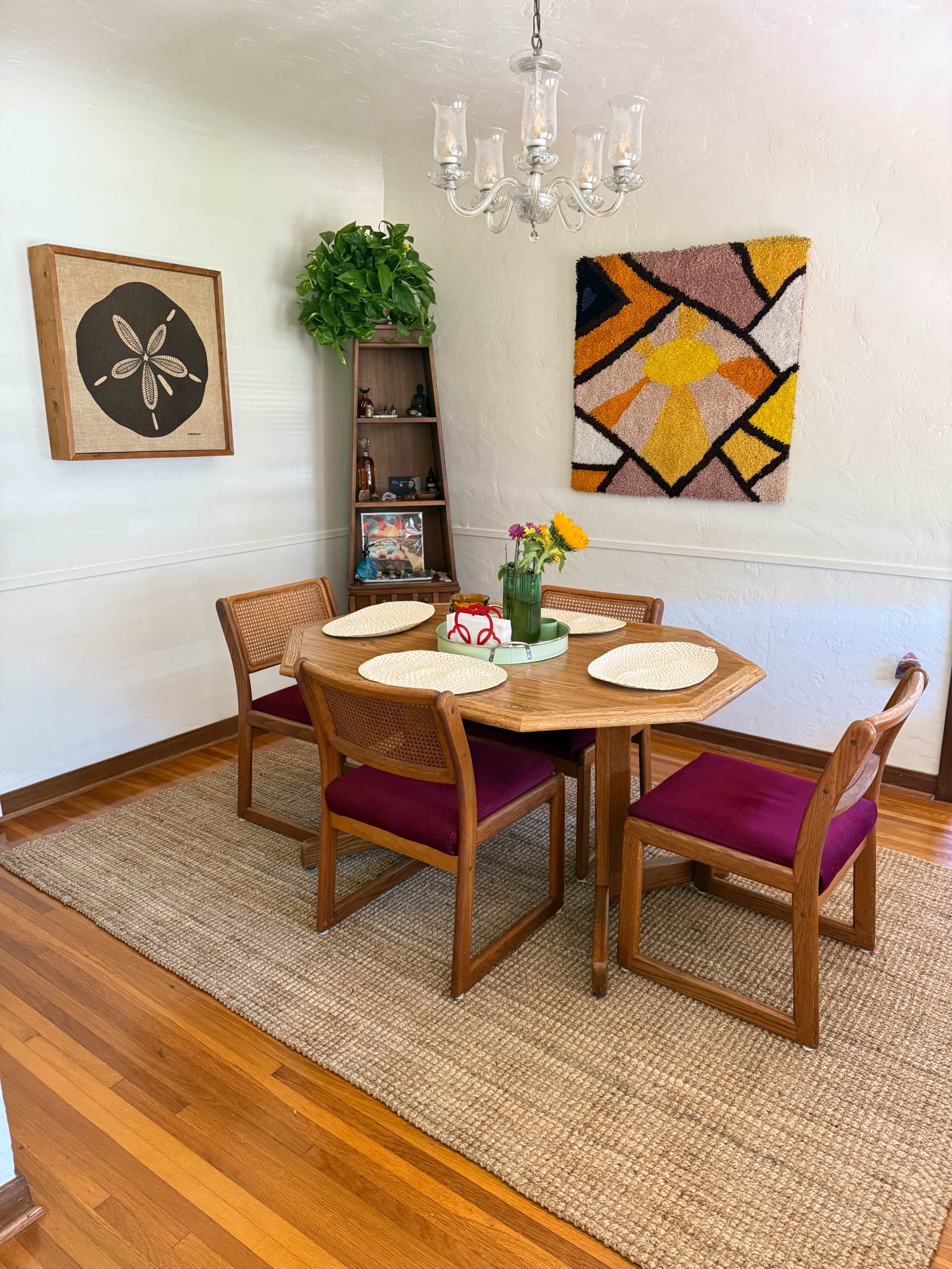 The image shows a dining area featuring a round wooden table with four chairs, a vase of flowers on the table, and a colorful wall hanging above a wooden shelf.