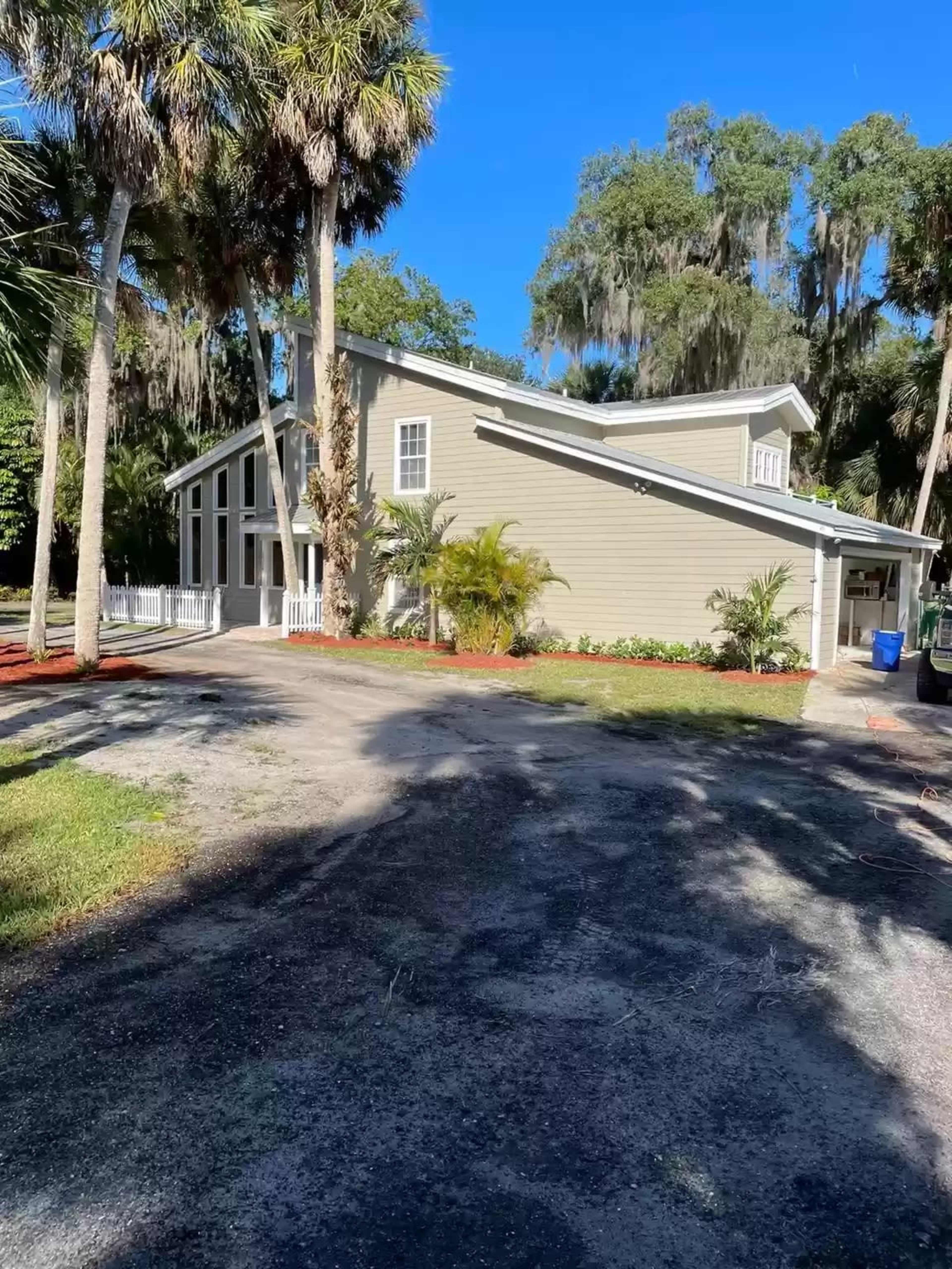 A two-story house with light-colored siding is situated among palm trees and surrounded by a landscaped yard.