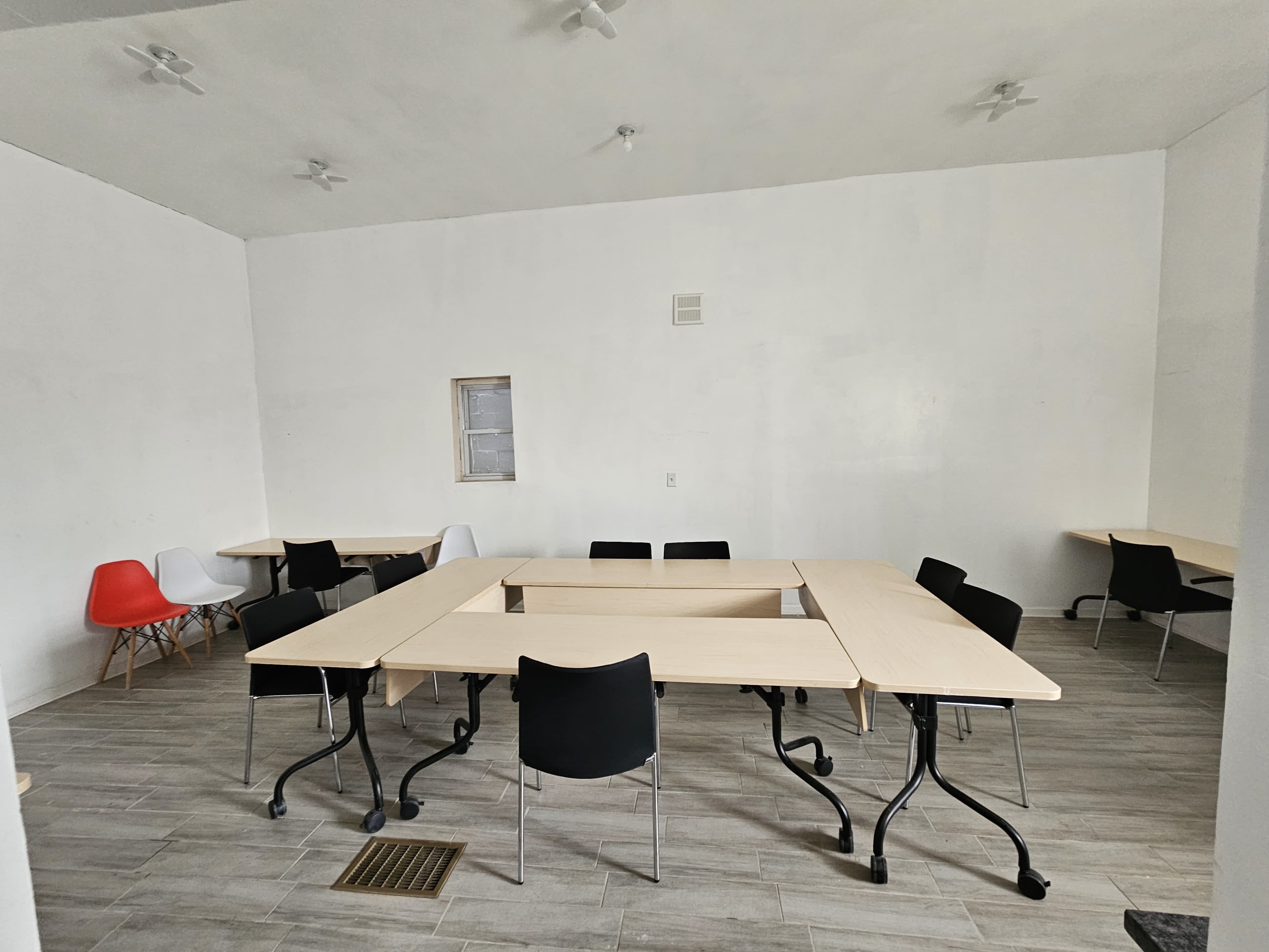 The image shows a spacious, white-walled room with several tables arranged in a square formation and additional chairs scattered around.