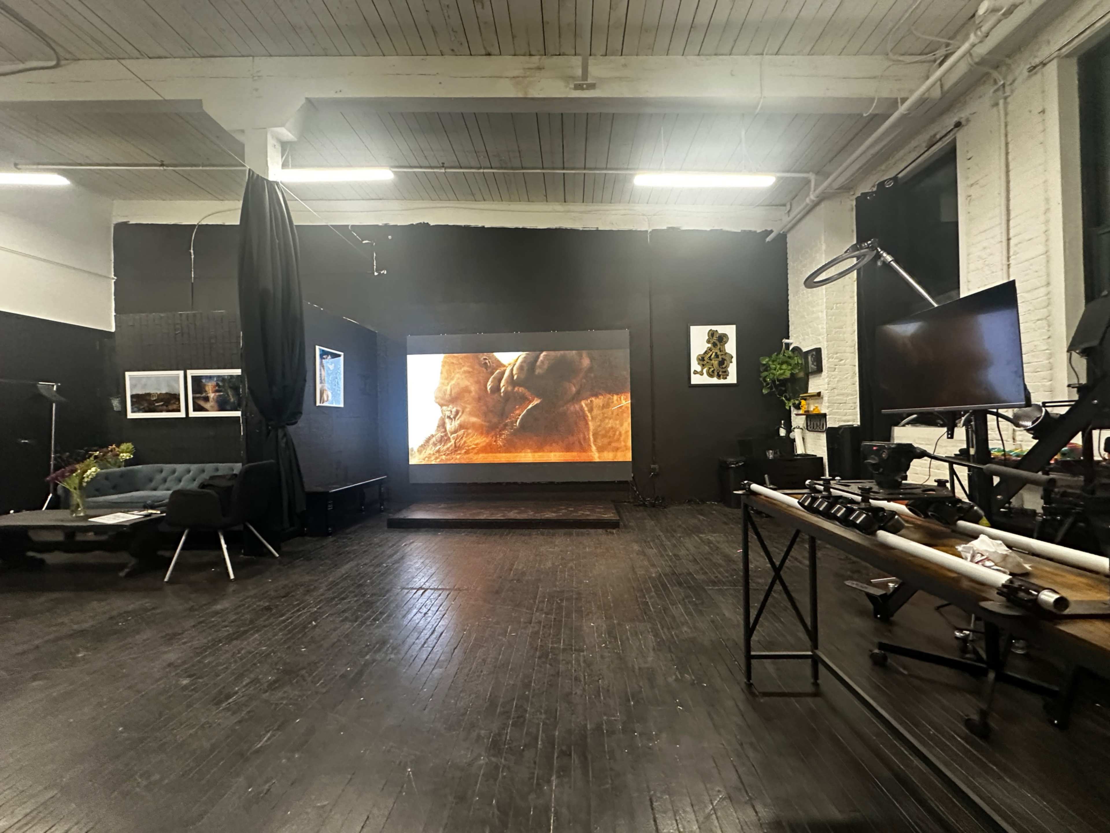 The image shows a spacious interior of a studio with a large projection screen displaying a vivid image, surrounded by furniture and artwork on the walls.