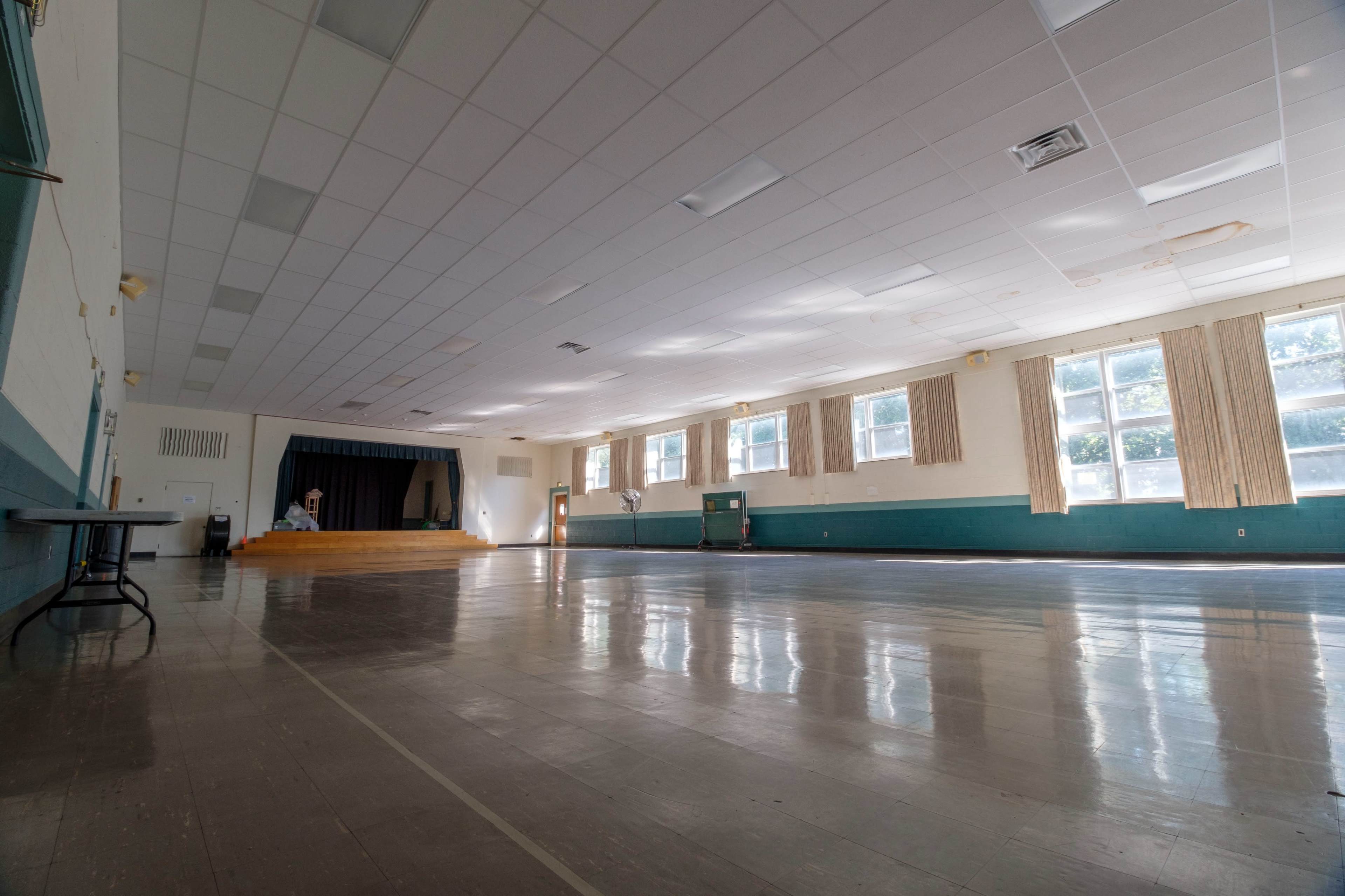 A spacious, empty hall with a stage at one end and large windows along the walls.