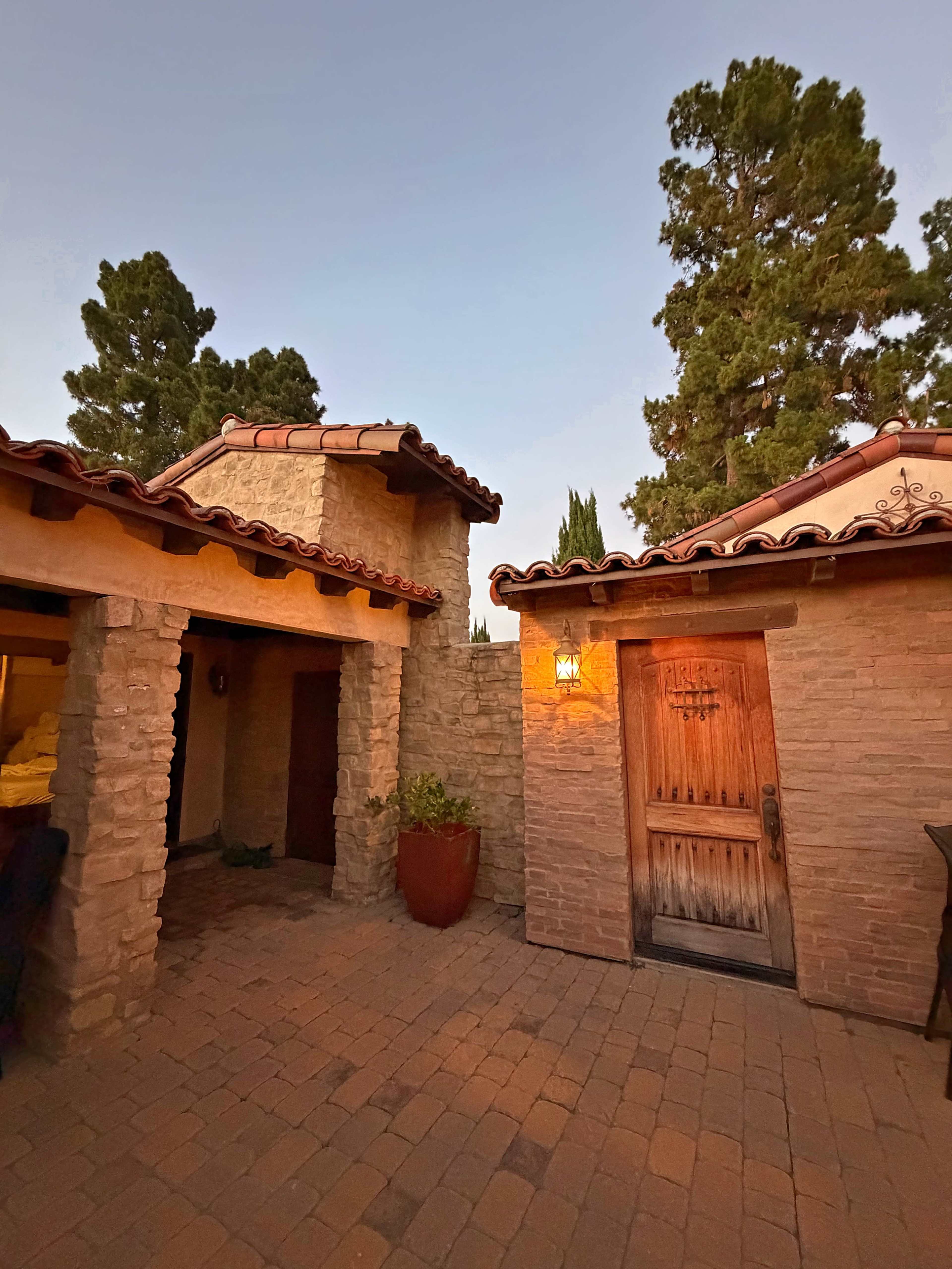 The image shows a stone and adobe-style exterior of a building, featuring a wooden door and a lantern, surrounded by tall green trees against a twilight sky.
