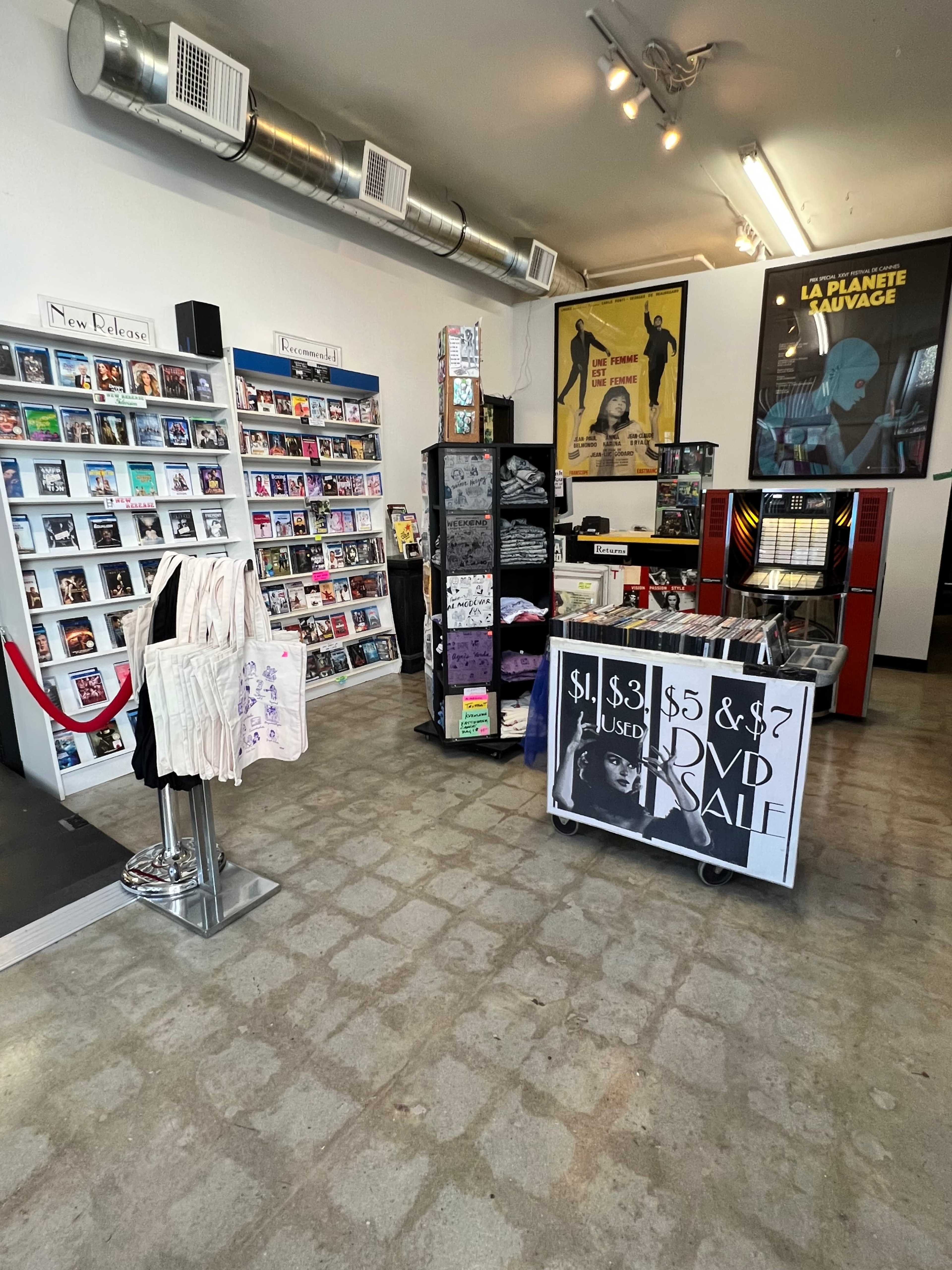 The interior of a retail store with shelves of DVDs and books, a display of merchandise, and a sale sign for used DVDs.
