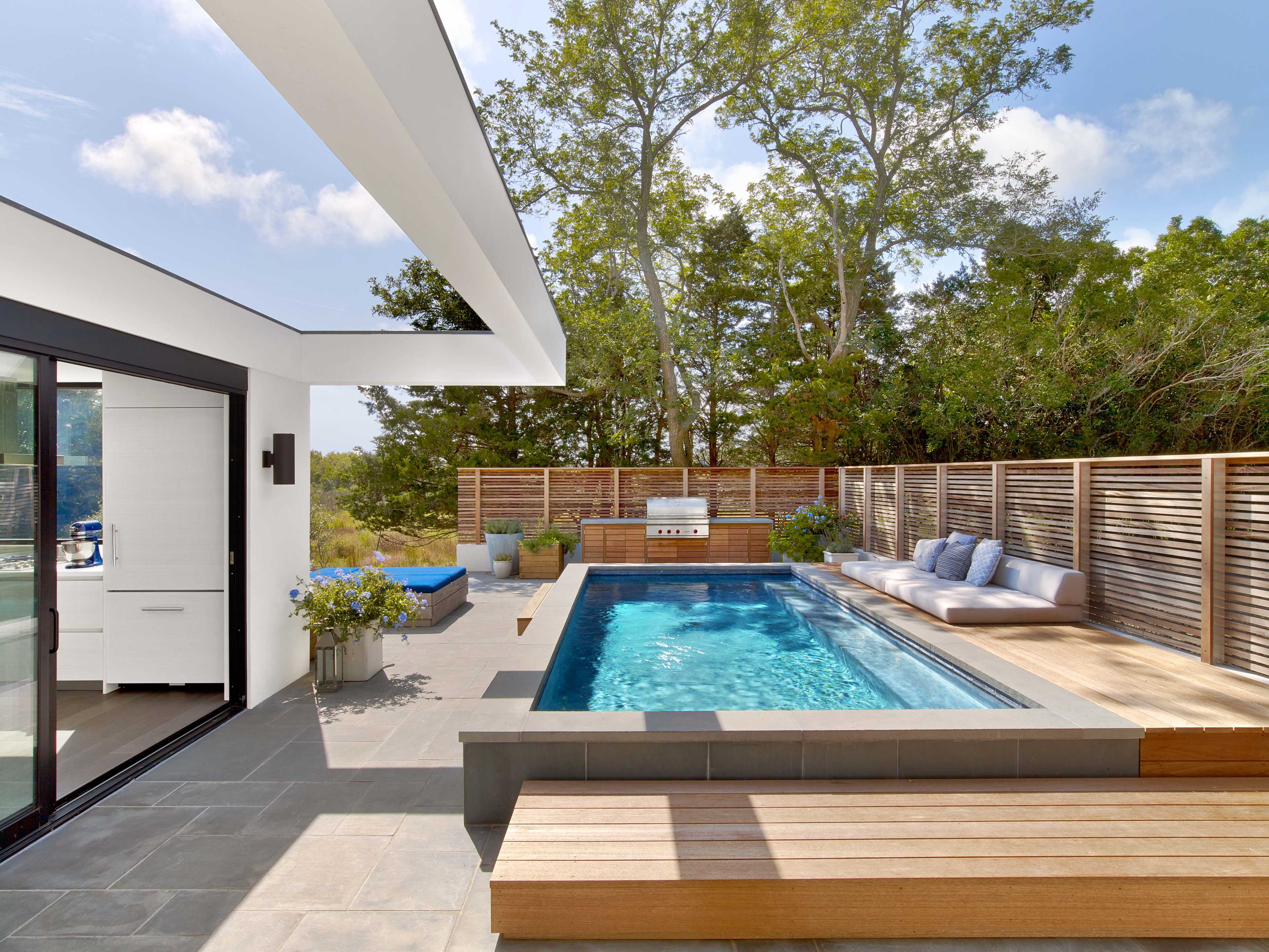 A modern outdoor space features a rectangular pool surrounded by a wooden deck, outdoor seating, and a barbecue area, set against a backdrop of trees.