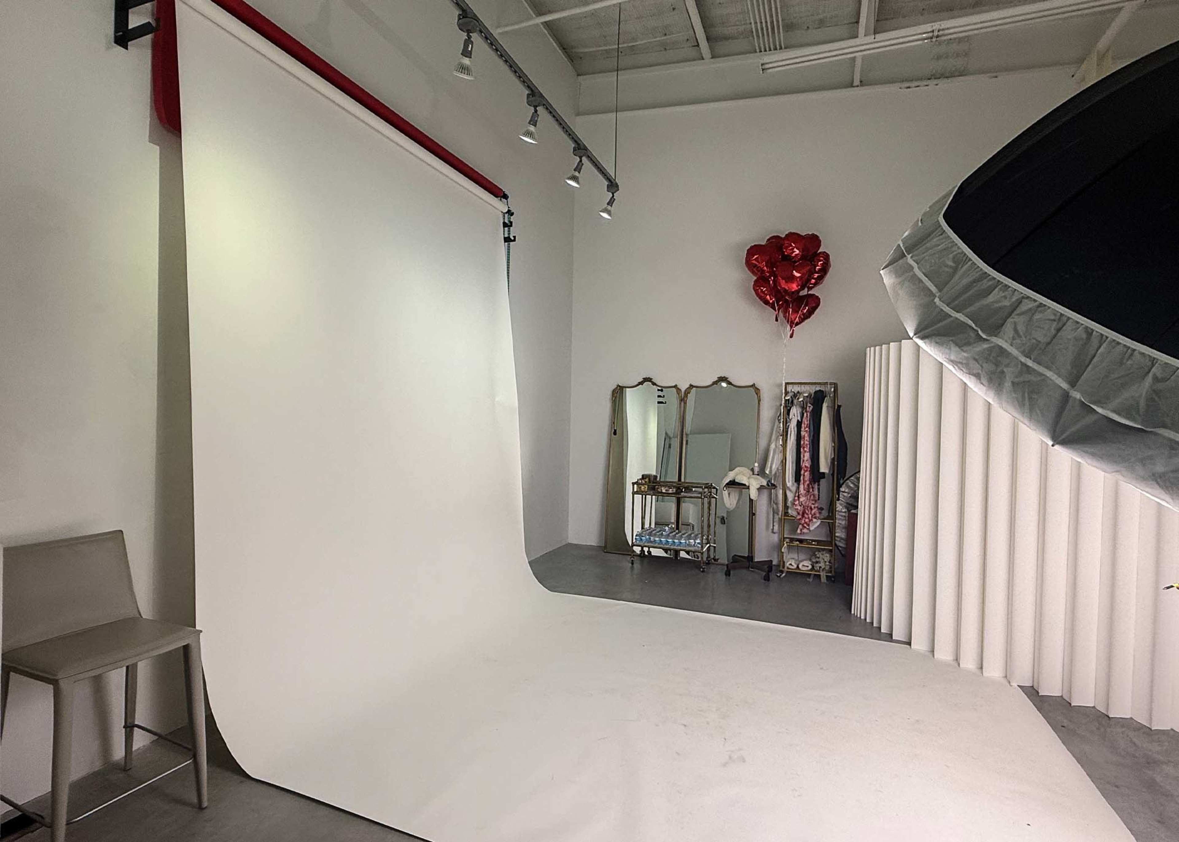 The image shows a photography studio with a white backdrop, a chair, a mirror, clothing racks, and heart-shaped balloons.
