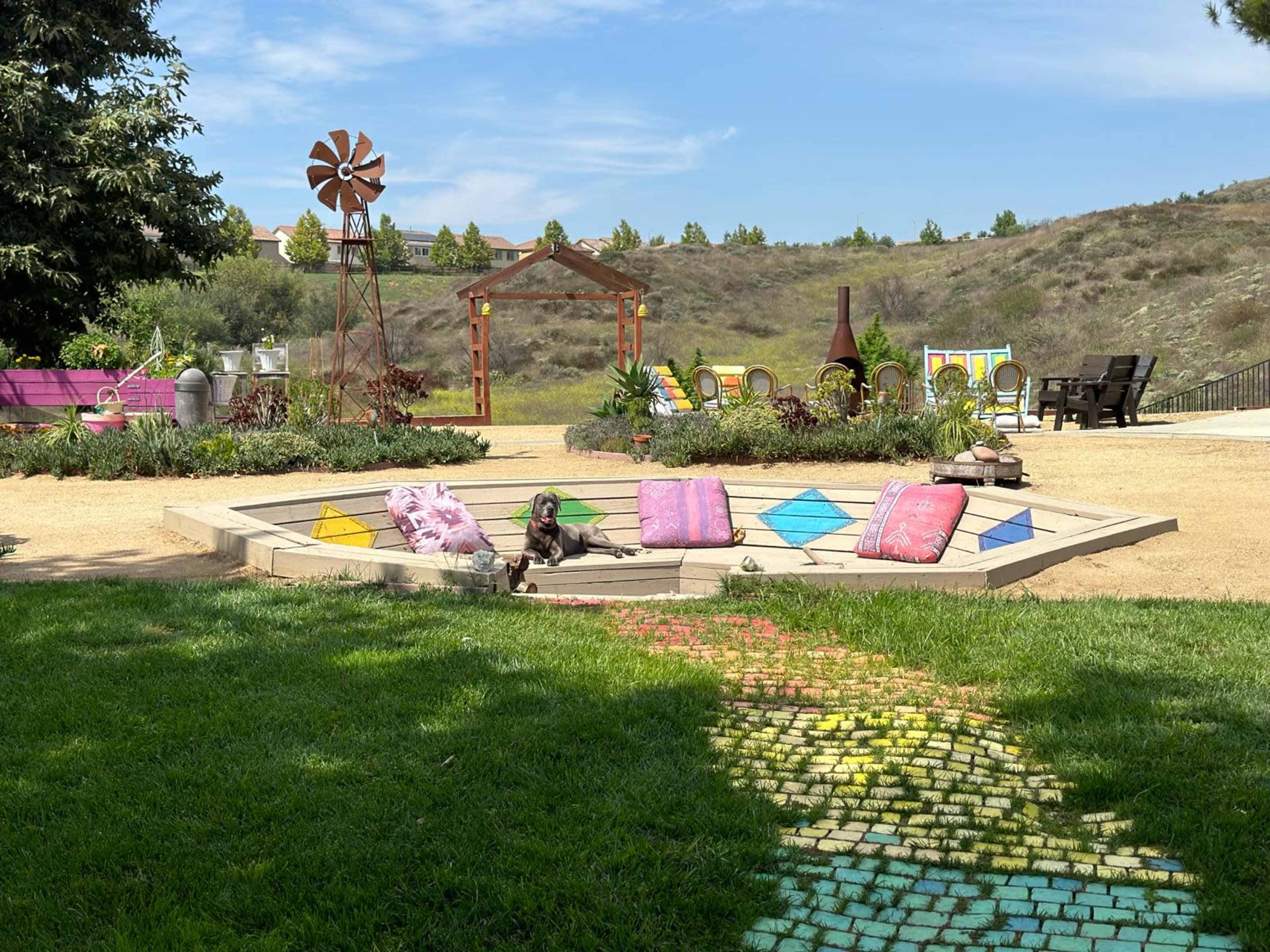 A landscaped backyard features a colorful pathway leading to a hexagonal seating area with cushions, surrounded by garden elements and a distant view of rolling hills.