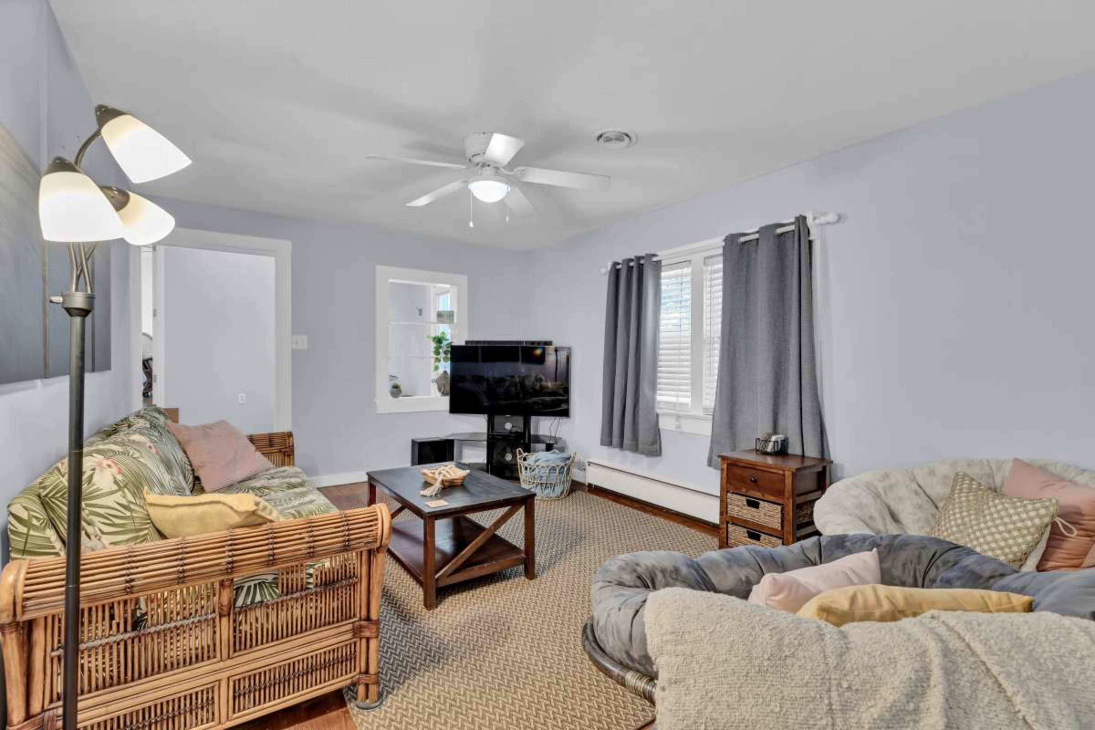 The image shows a living room featuring a comfortable seating area with a patterned couch and a large, cozy armchair, a coffee table, a television on a stand, and light-colored walls with curtains.