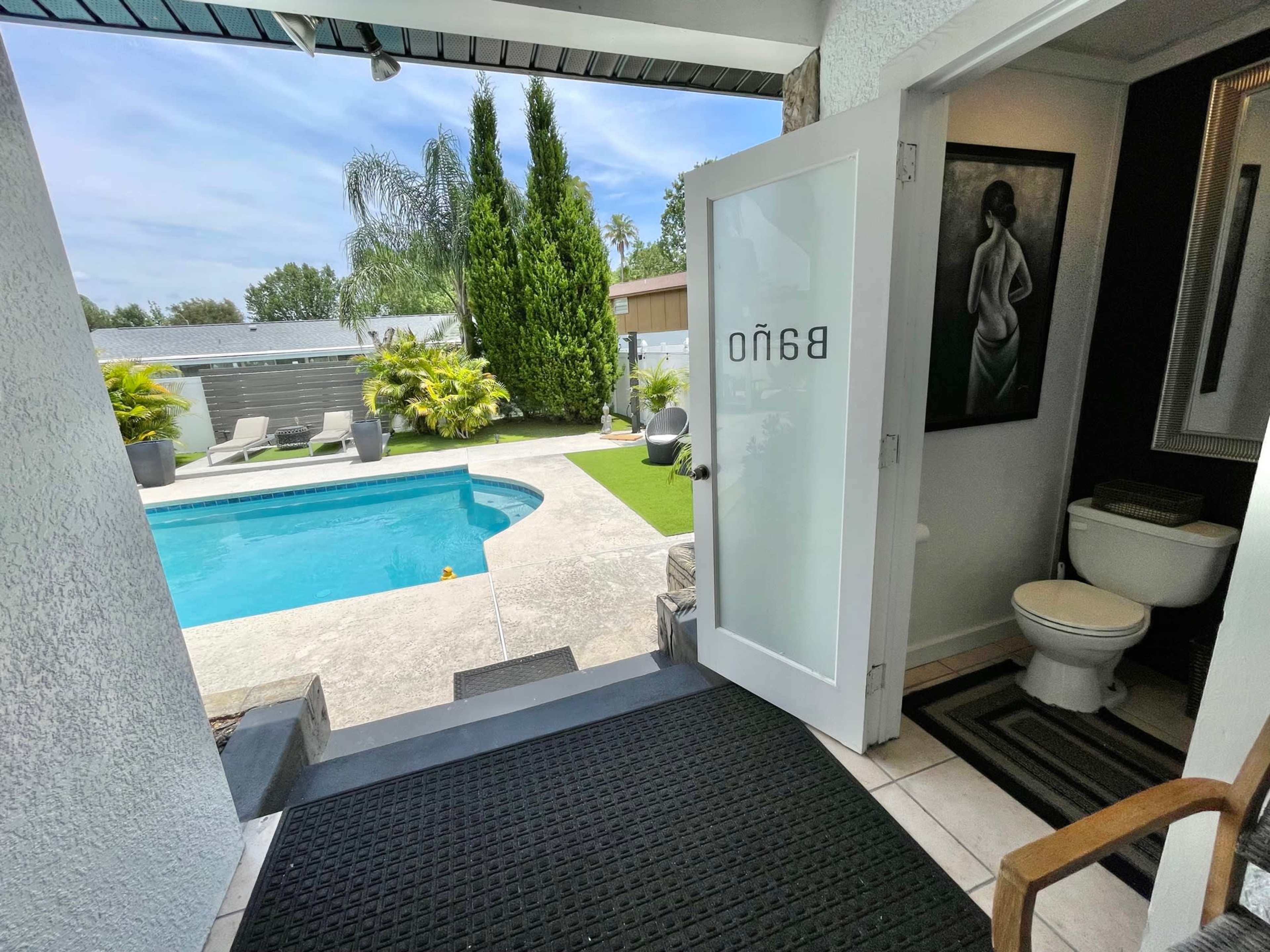 A view of a well-lit bathroom entrance leading to a swimming pool surrounded by greenery.