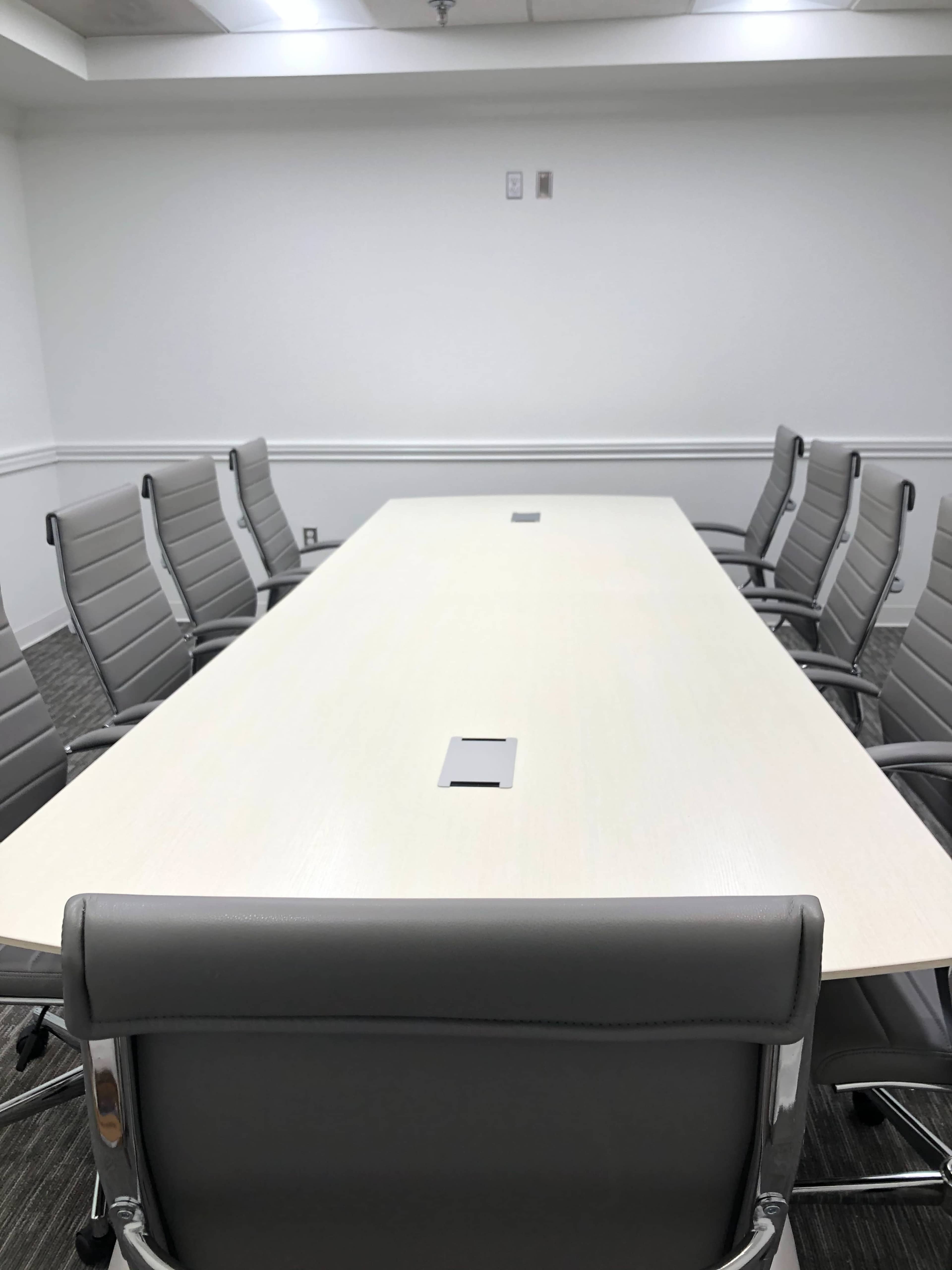 A long, light-colored table is surrounded by eight gray office chairs in a bright conference room.