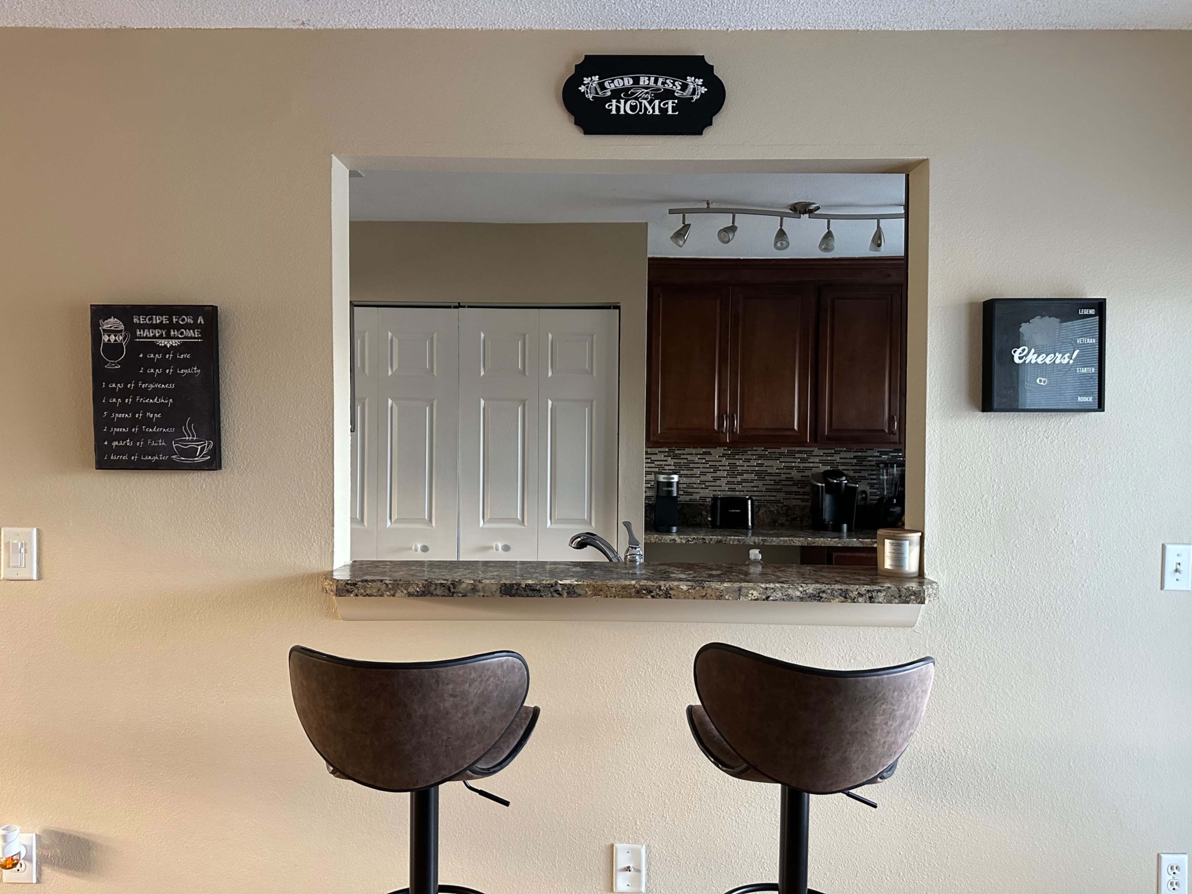 A bar area features two stools in front of a granite countertop with a wall-mounted sign above and cabinets in the background.
