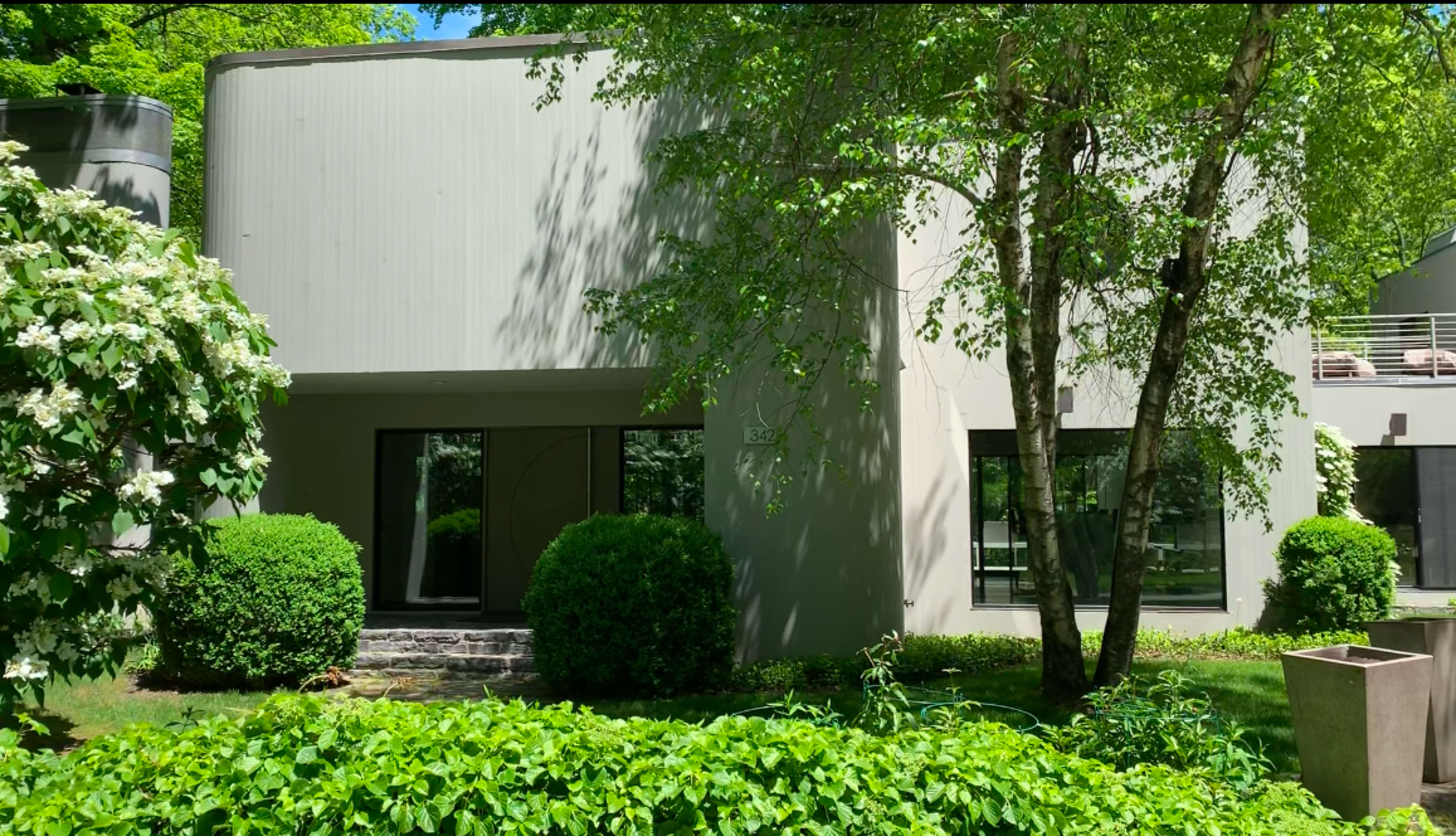 The image shows a modern, two-story house with large windows, surrounded by neatly trimmed bushes and trees in a green landscape.