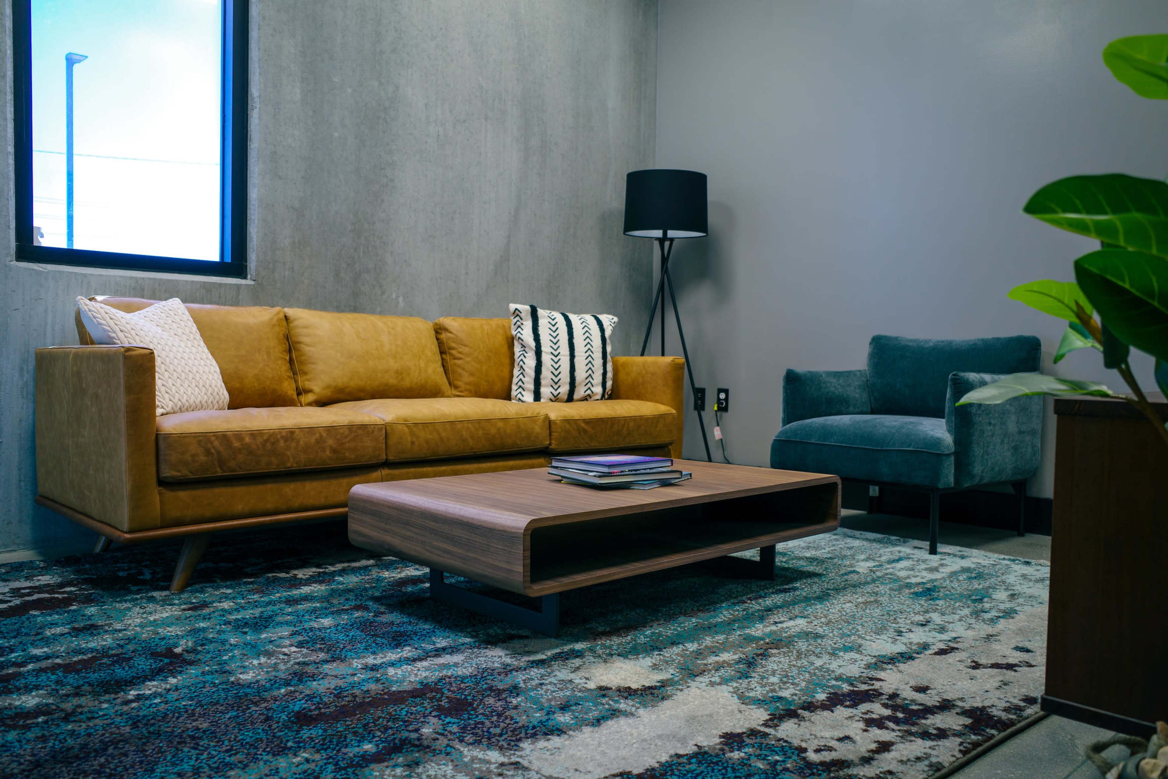 The image shows a modern living area with a brown leather sofa, a blue armchair, a wooden coffee table, and a patterned rug on a concrete floor.