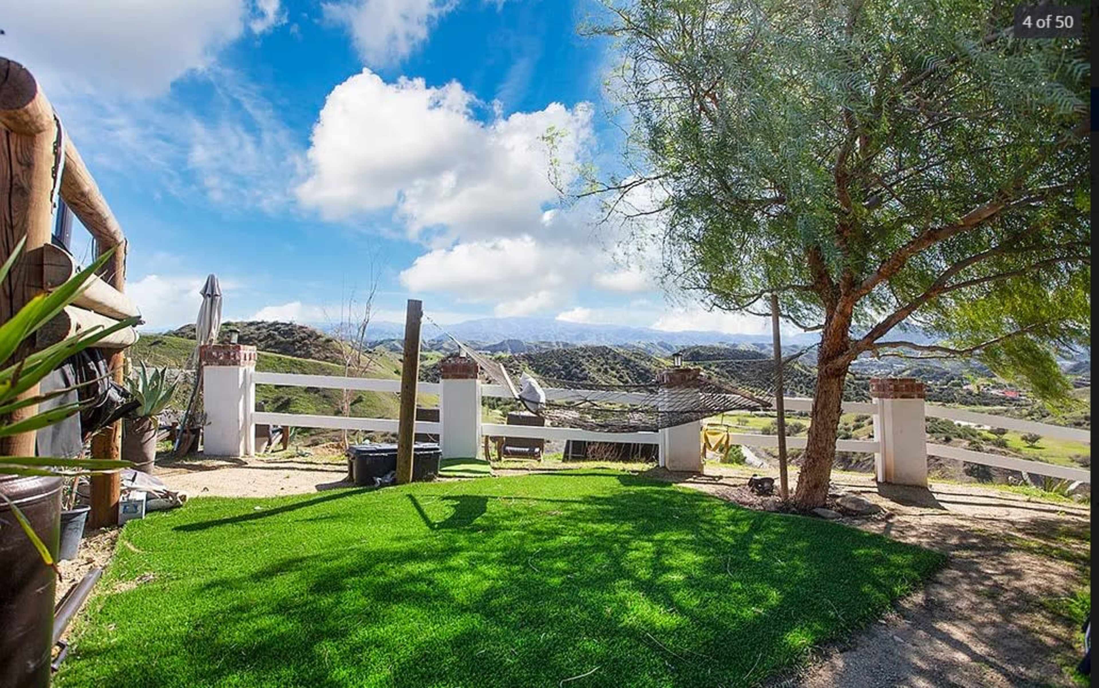 A landscaped area with artificial turf and a tree overlooks a hilly landscape under a partly cloudy sky.