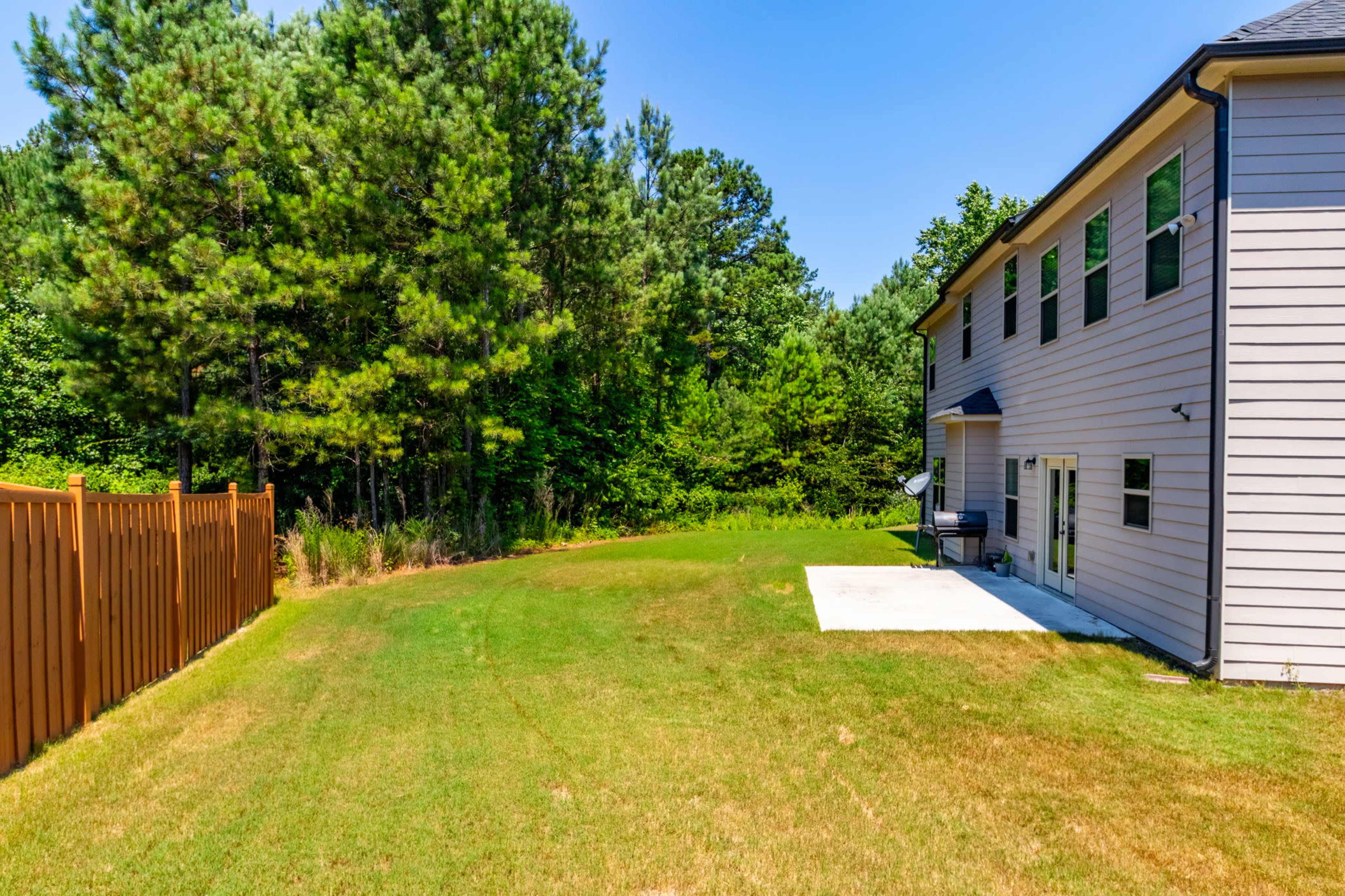 A neatly maintained backyard features a grassy area adjacent to a house and a wooden fence, with trees forming a natural border in the background.