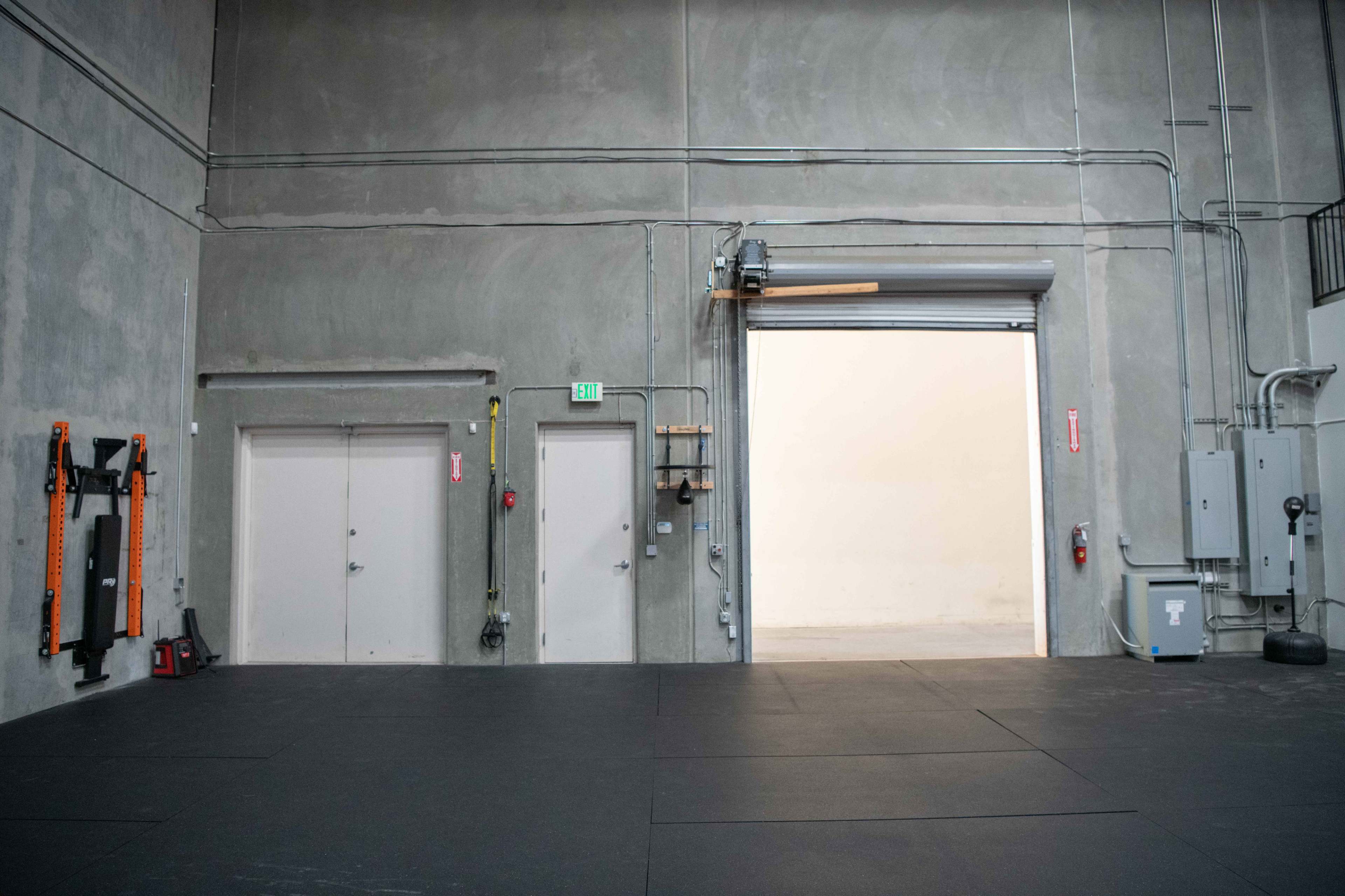An industrial space featuring a gray wall, a large open door, two smaller doors, and various electrical equipment along the walls.