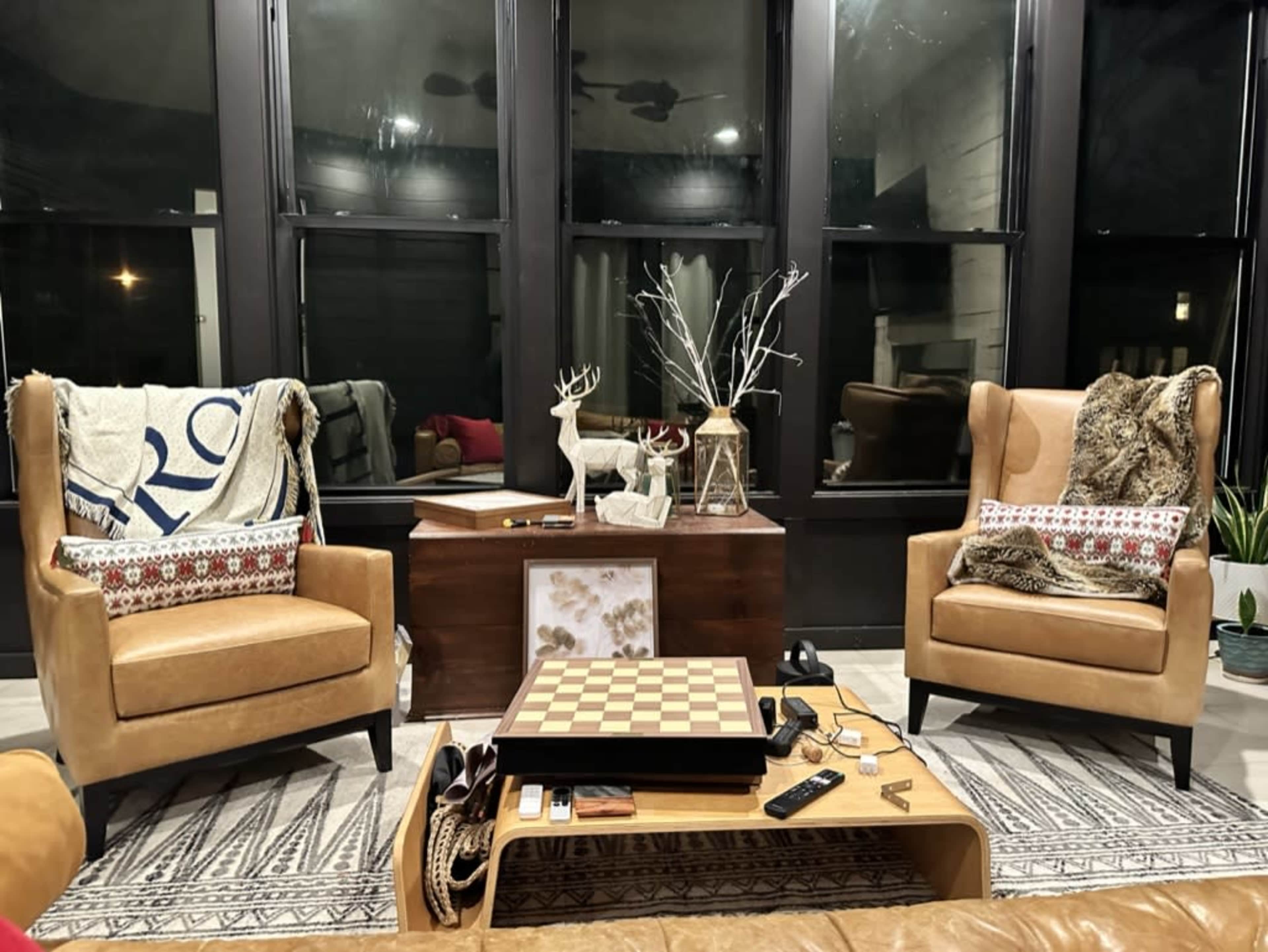 A cozy living space with two tan chairs facing a coffee table, a chess board on the table, and decorative elements like a plant and an antler display in the background.