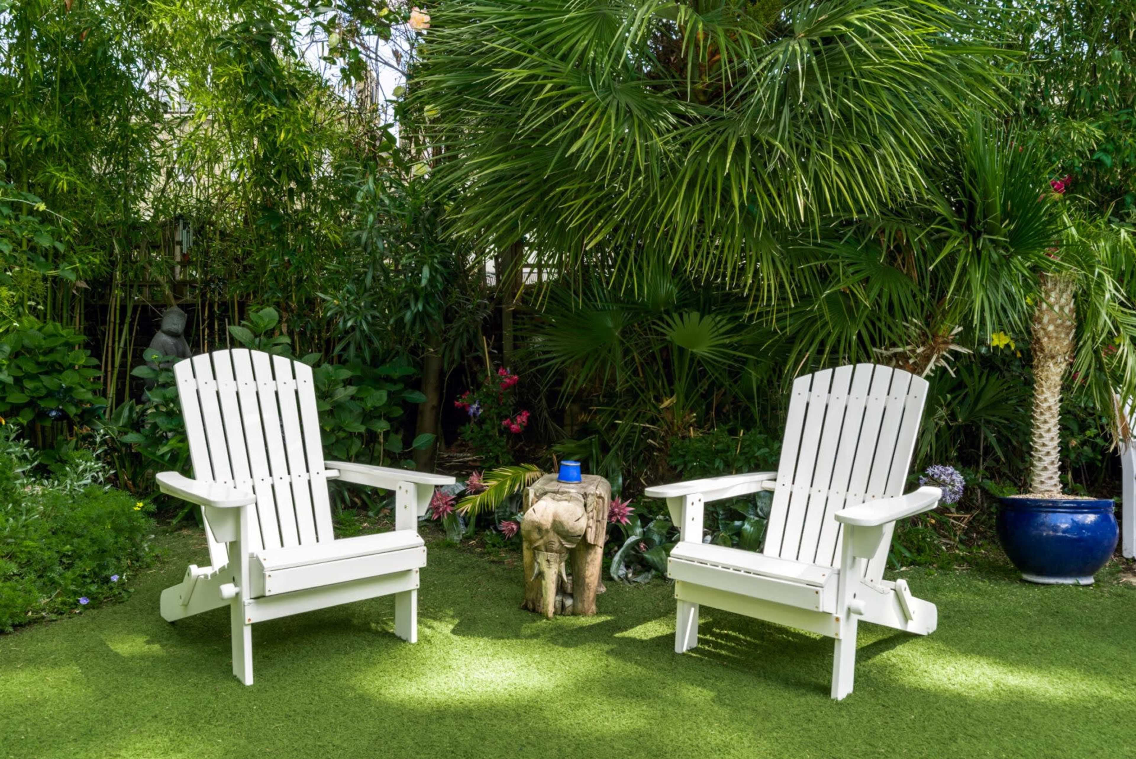 Two white Adirondack chairs sit on a grassy area surrounded by lush greenery and tropical plants.