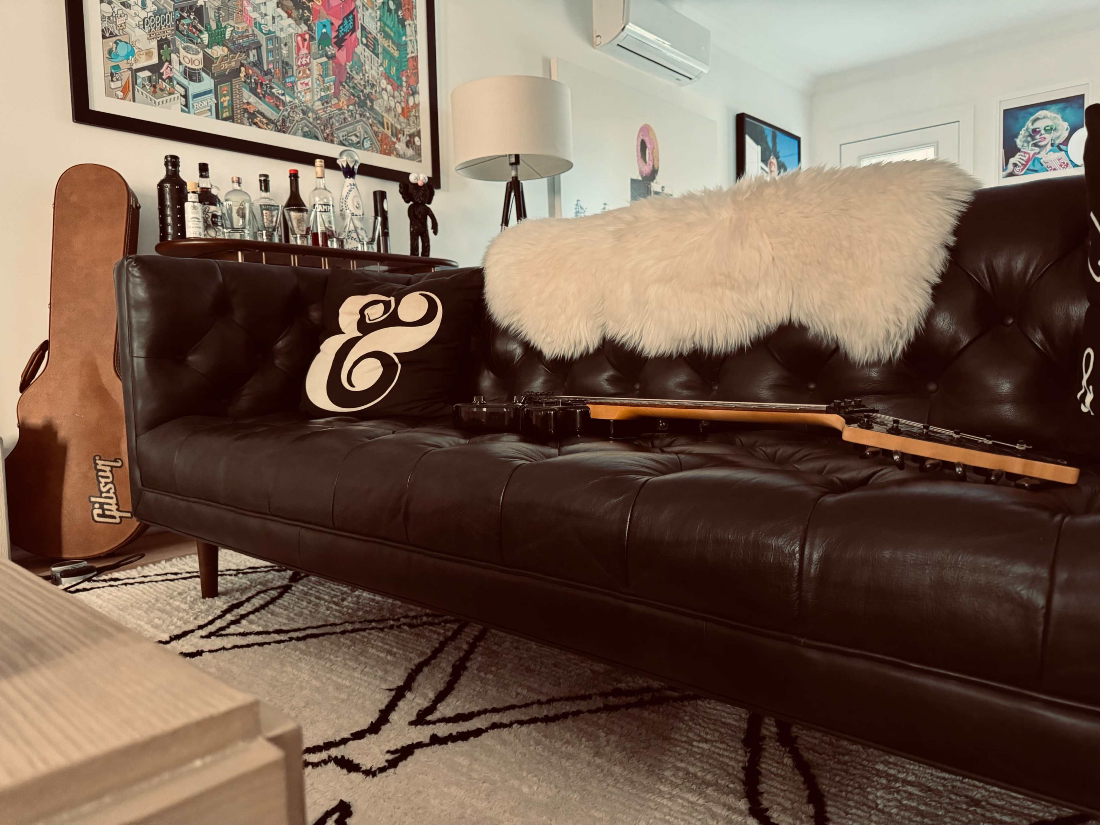 A black leather sofa with a white throw and a decorative cushion sits in a stylish living room, alongside a guitar and a collection of bottles on a shelf.