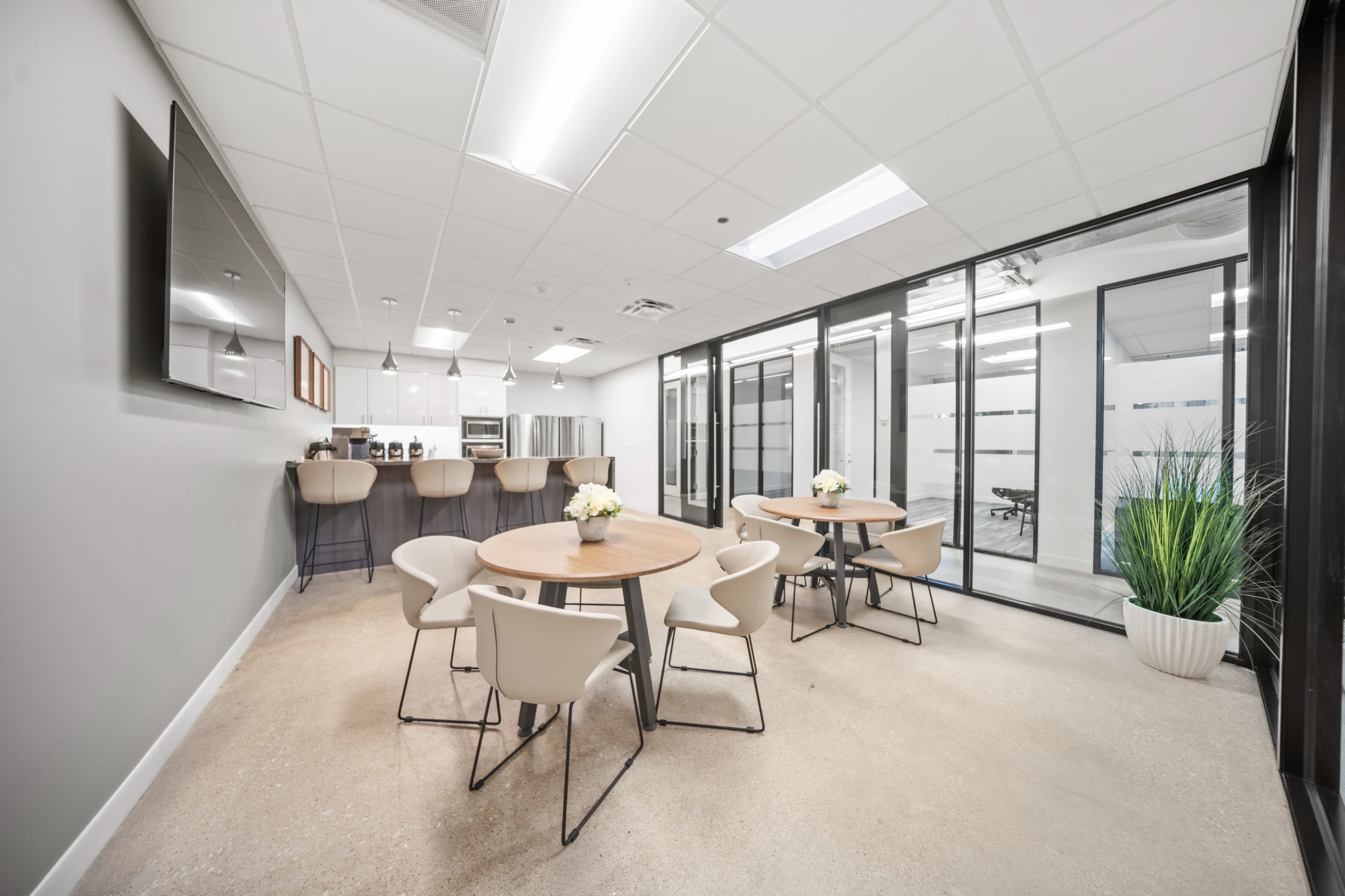 The image shows a modern office break room featuring two round tables with chairs, a kitchenette area with appliances, and glass-walled meeting rooms.
