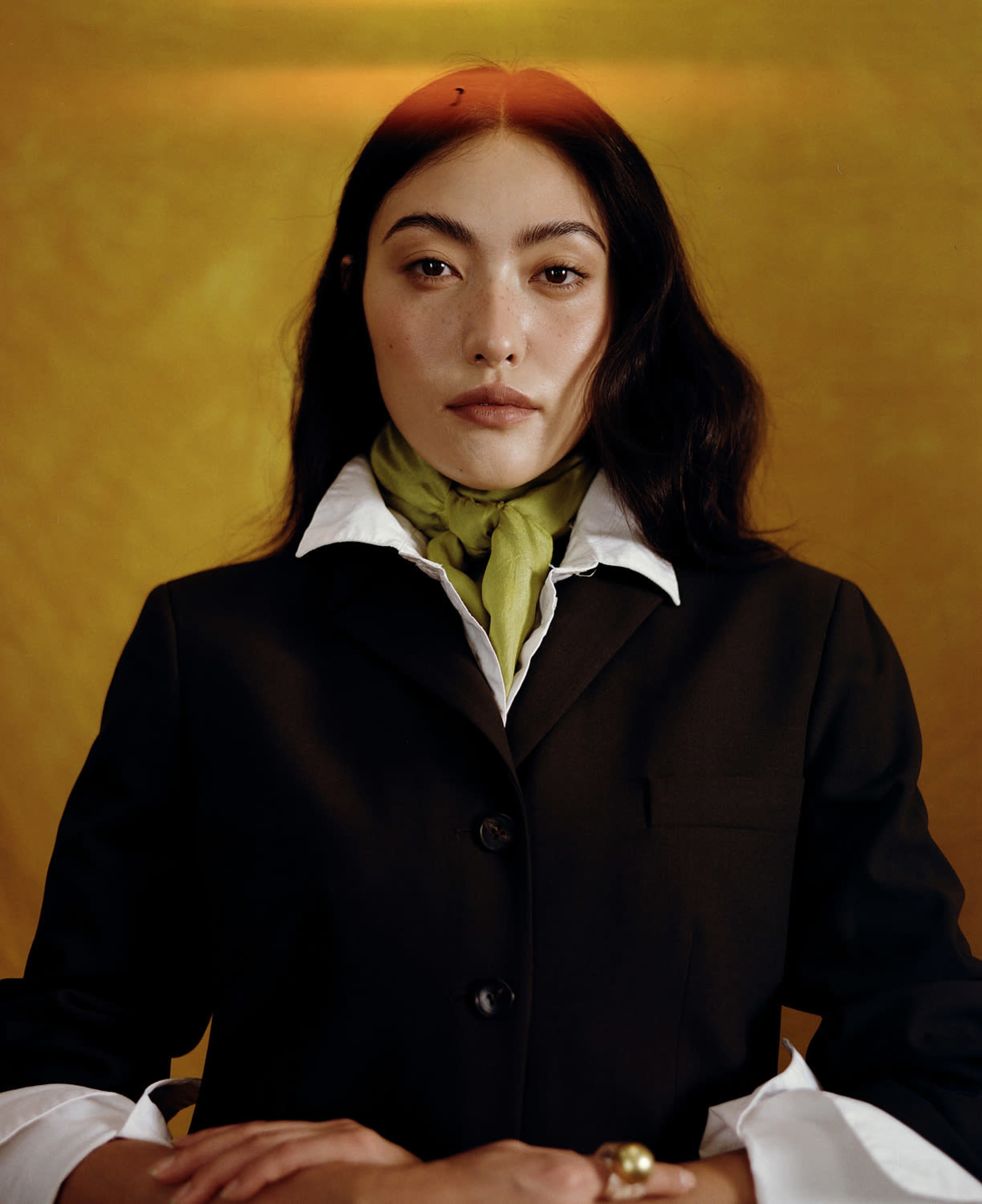 A woman in a black blazer and white shirt, wearing a green scarf, poses against a yellow background.