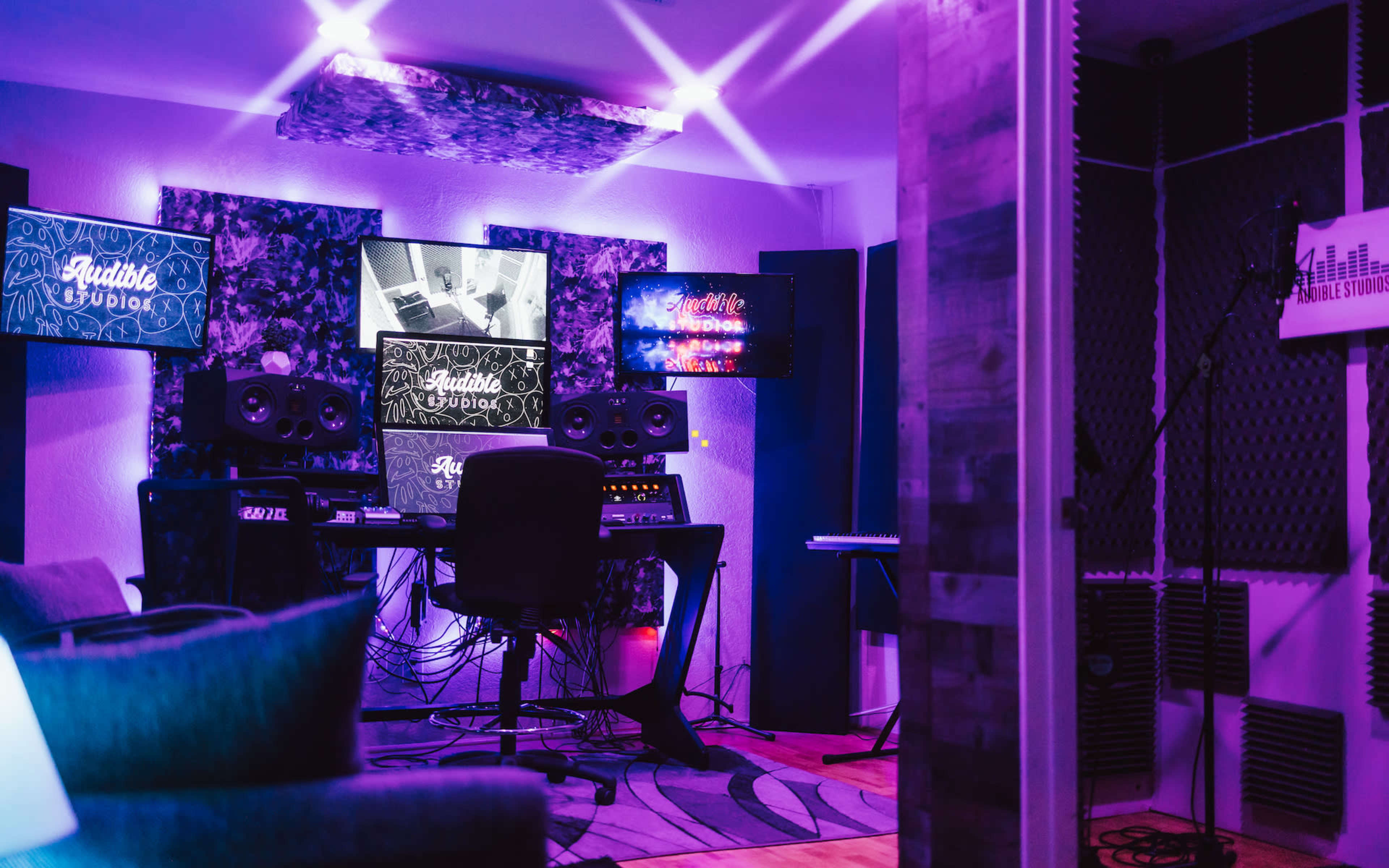 A music production studio features a central workstation with monitors, speakers, and equipment, surrounded by colorful lights and soundproofing materials.