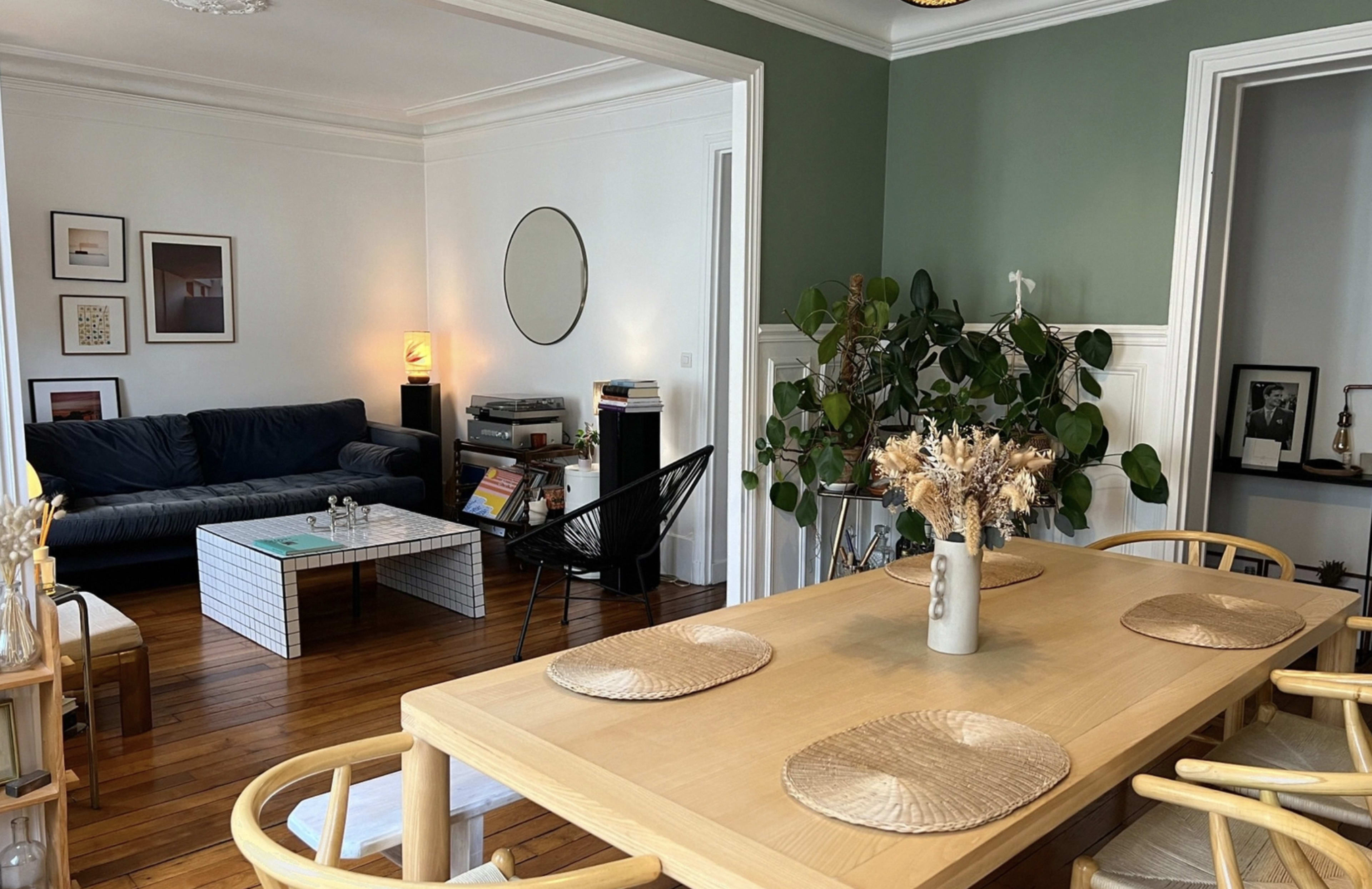 The image shows a well-decorated living and dining area featuring a plush sofa, a round mirror, a dining table with decorative place settings, and an assortment of indoor plants.