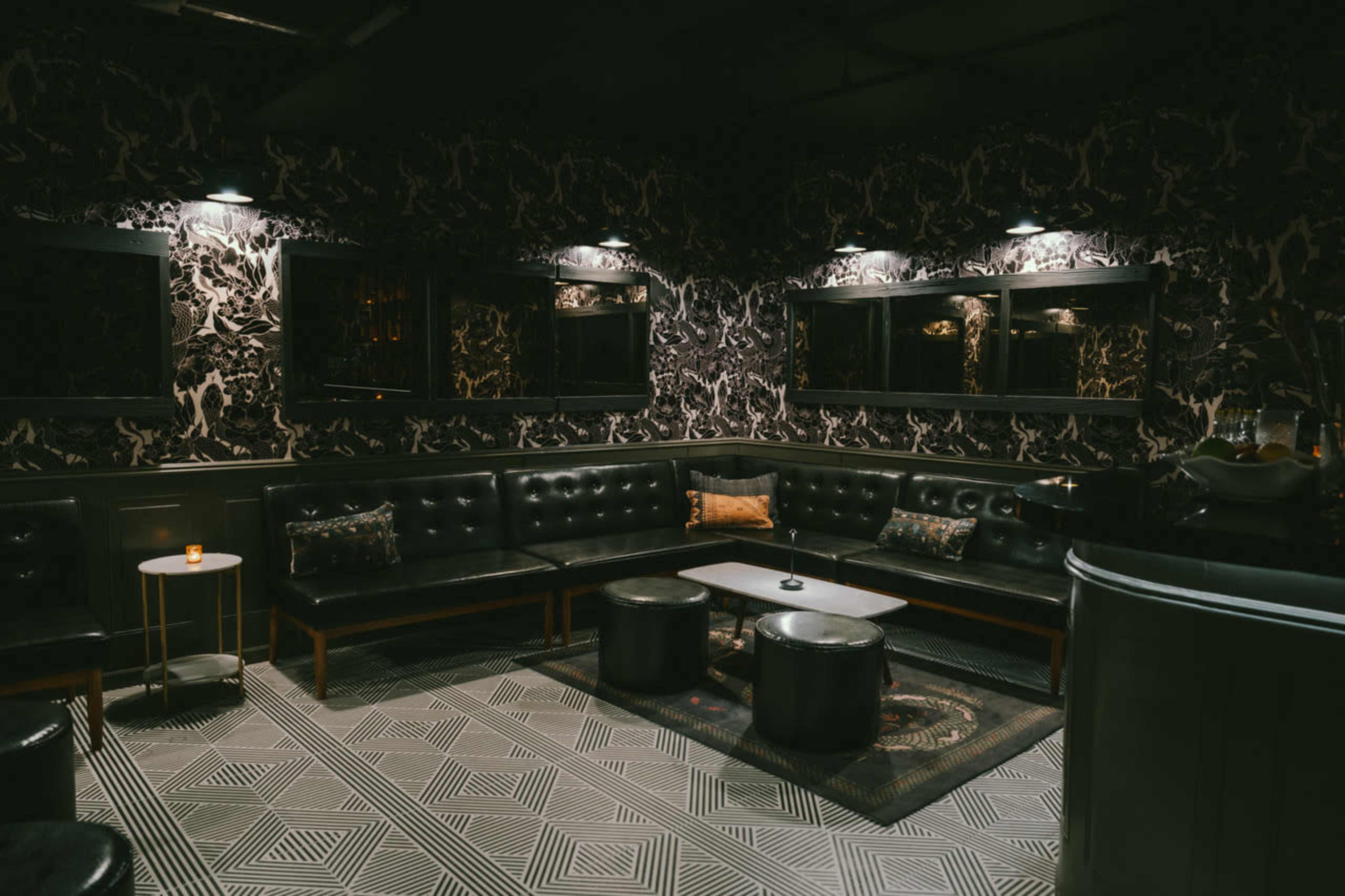 The image shows a dimly lit lounge area with black leather seating, patterned wallpaper, and a geometric tiled floor.