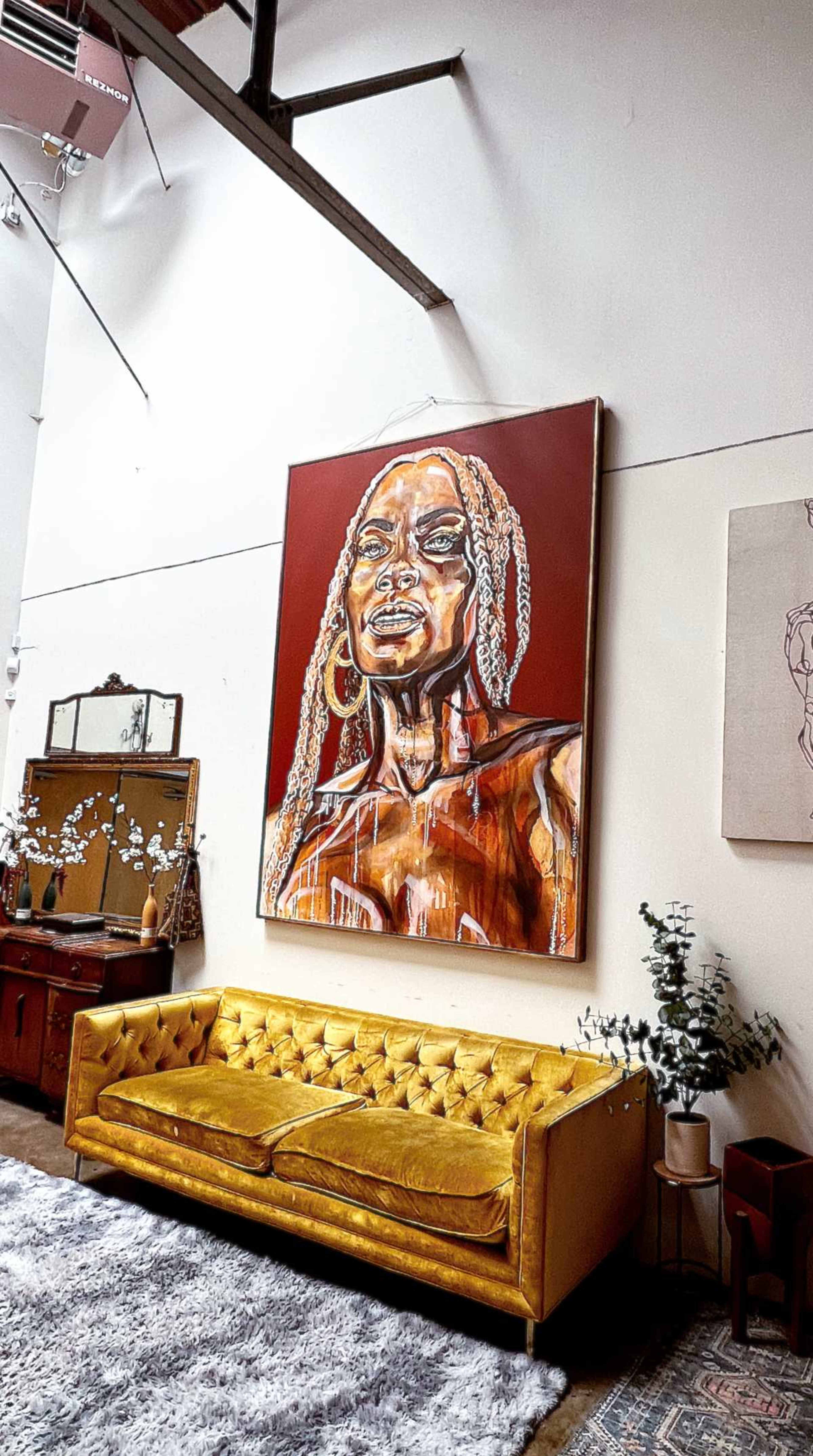 A large painting of a woman hangs on the wall above a gold tufted sofa in a bright, open interior space.
