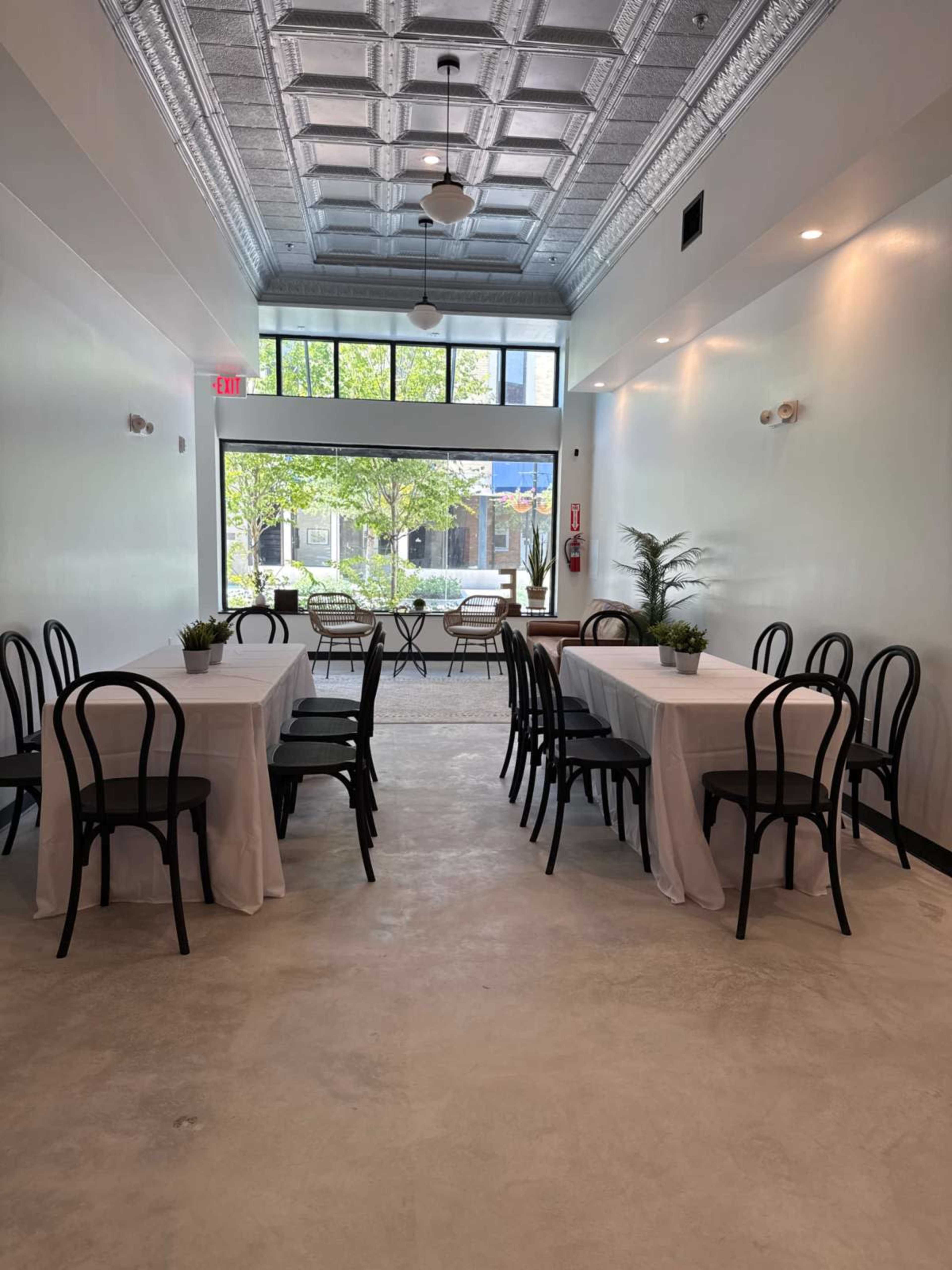 The image shows a bright, empty dining area with two rows of tables covered in white tablecloths and large windows providing natural light.
