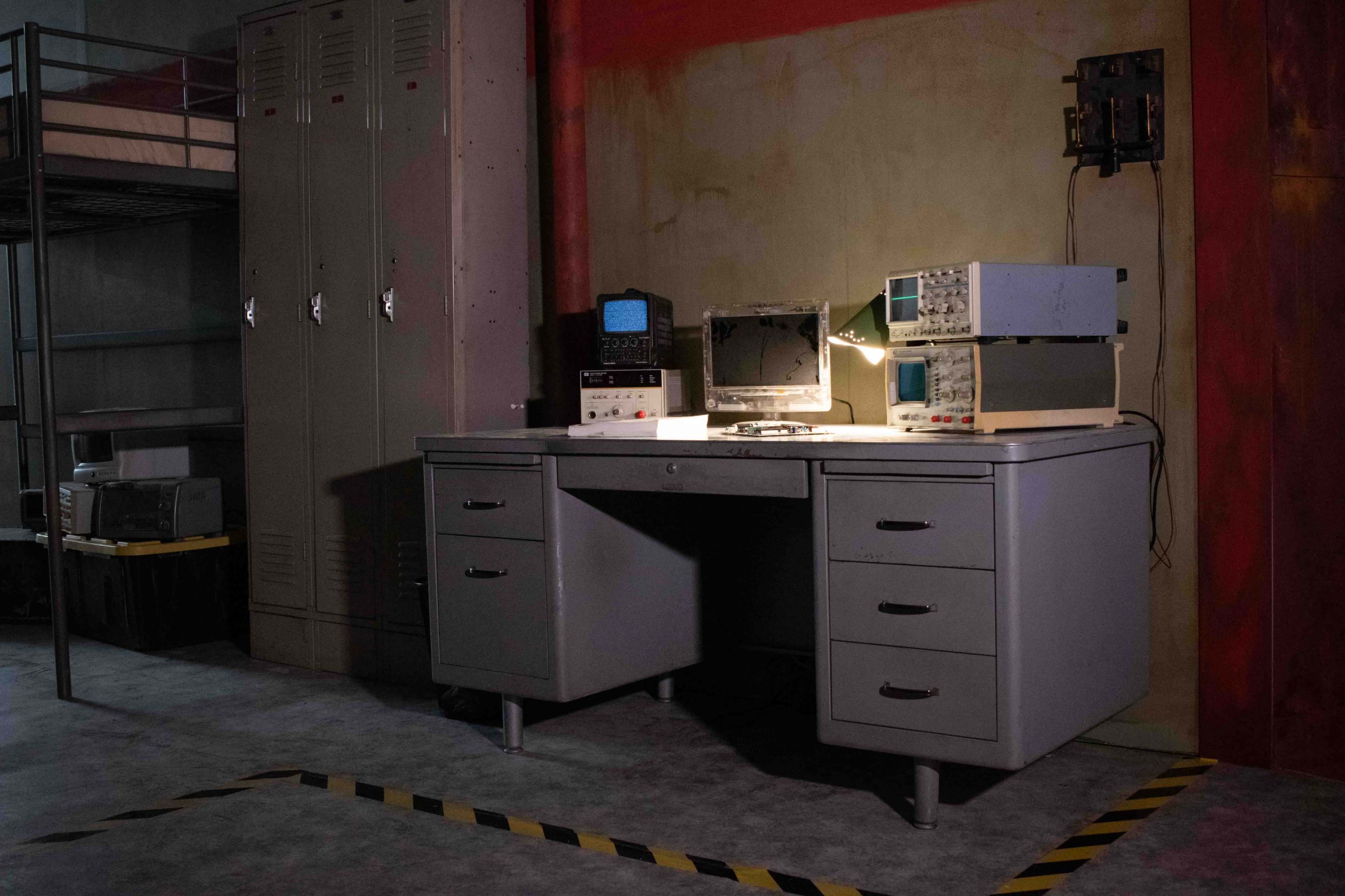 A gray metal desk with drawers is positioned next to lockers, with electronic equipment and a lamp on it in a dimly lit room.