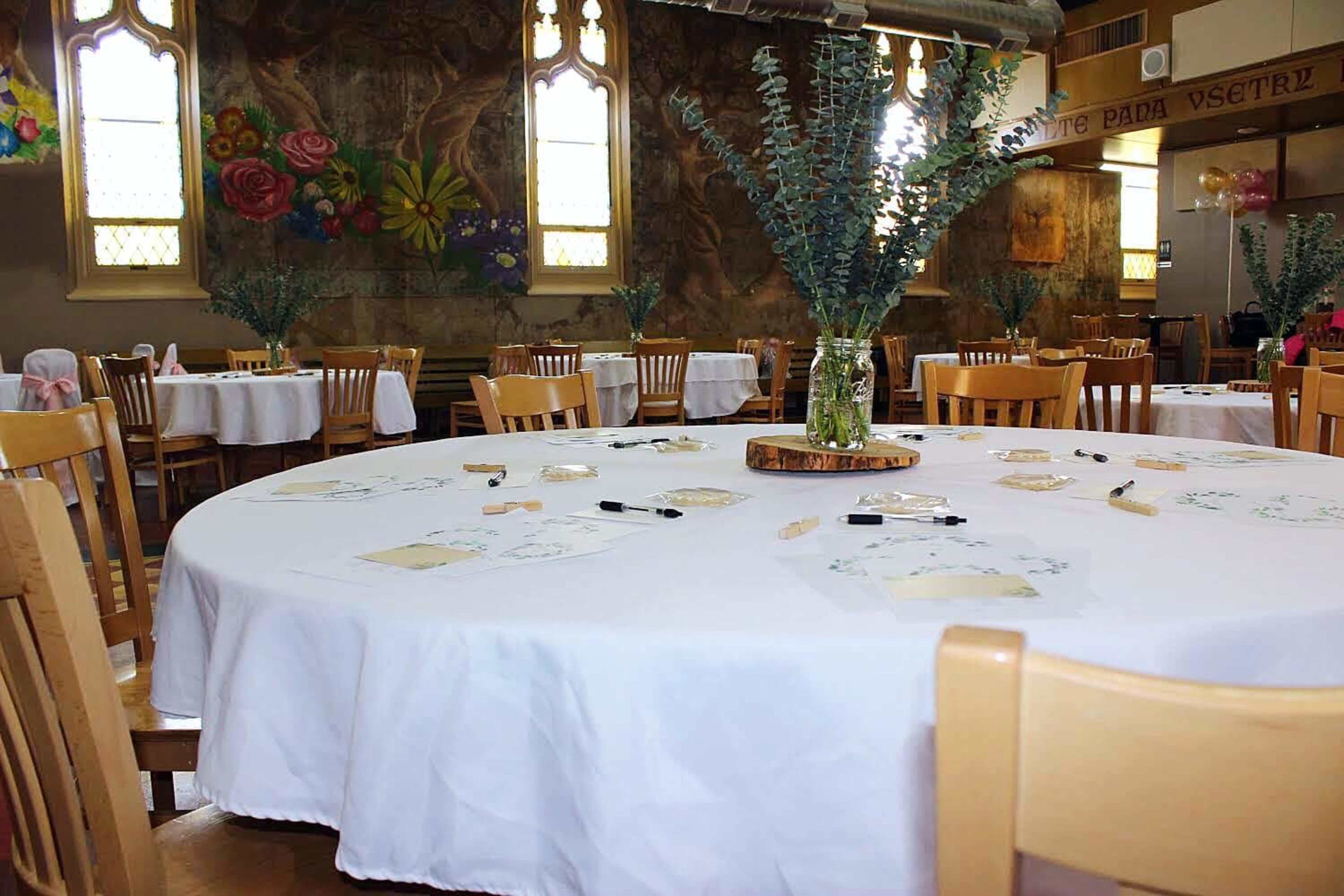 A dining area set with round tables covered in white tablecloths, decorated with vases of foliage and situated near a floral mural.