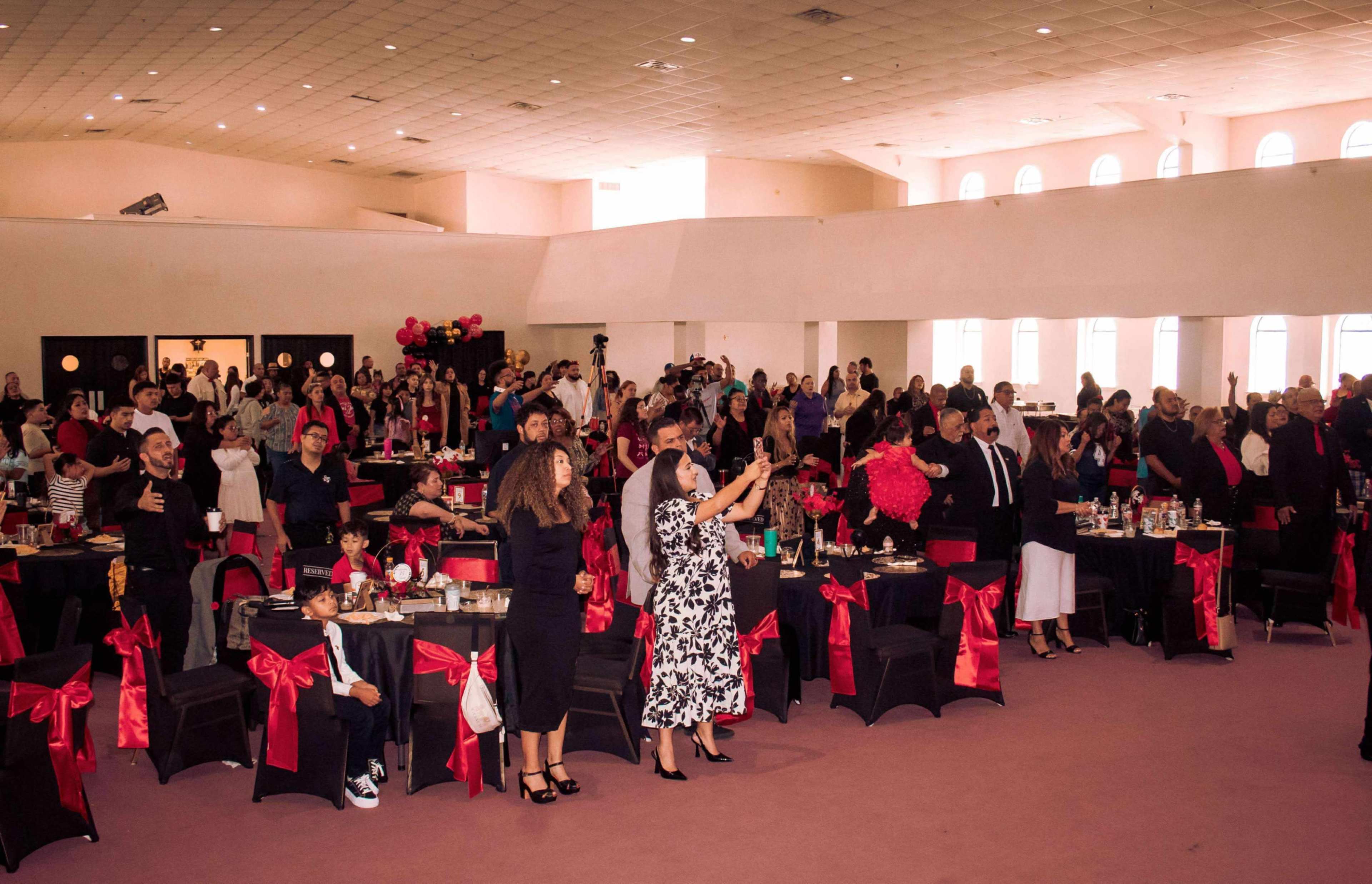 A large crowd gathers in a hall decorated with tables and chairs covered in black cloth and red bows, as they engage in a celebratory event.