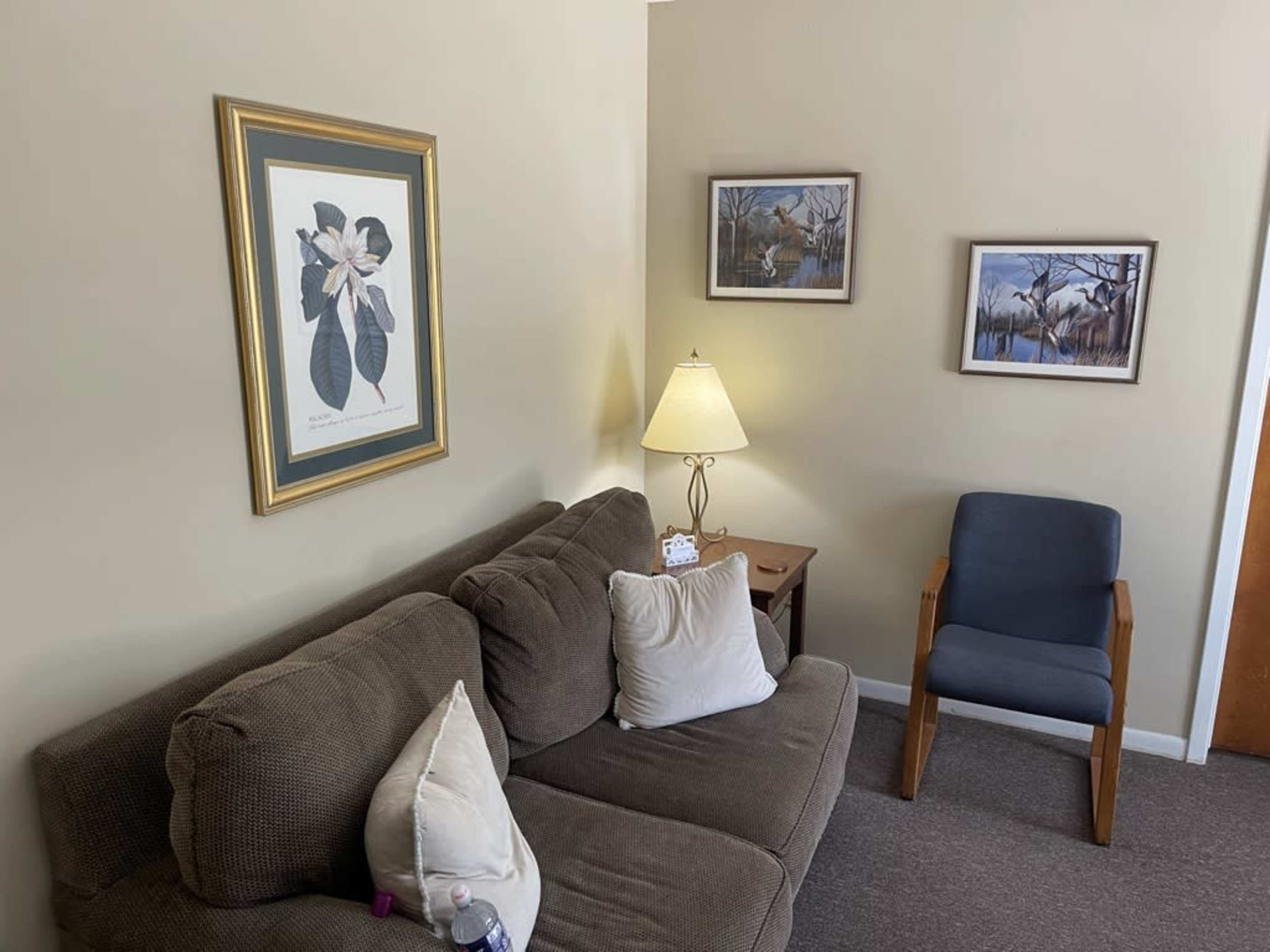 A small living area with a brown sofa, a lamp on a side table, two framed pictures on the wall, and a blue chair.