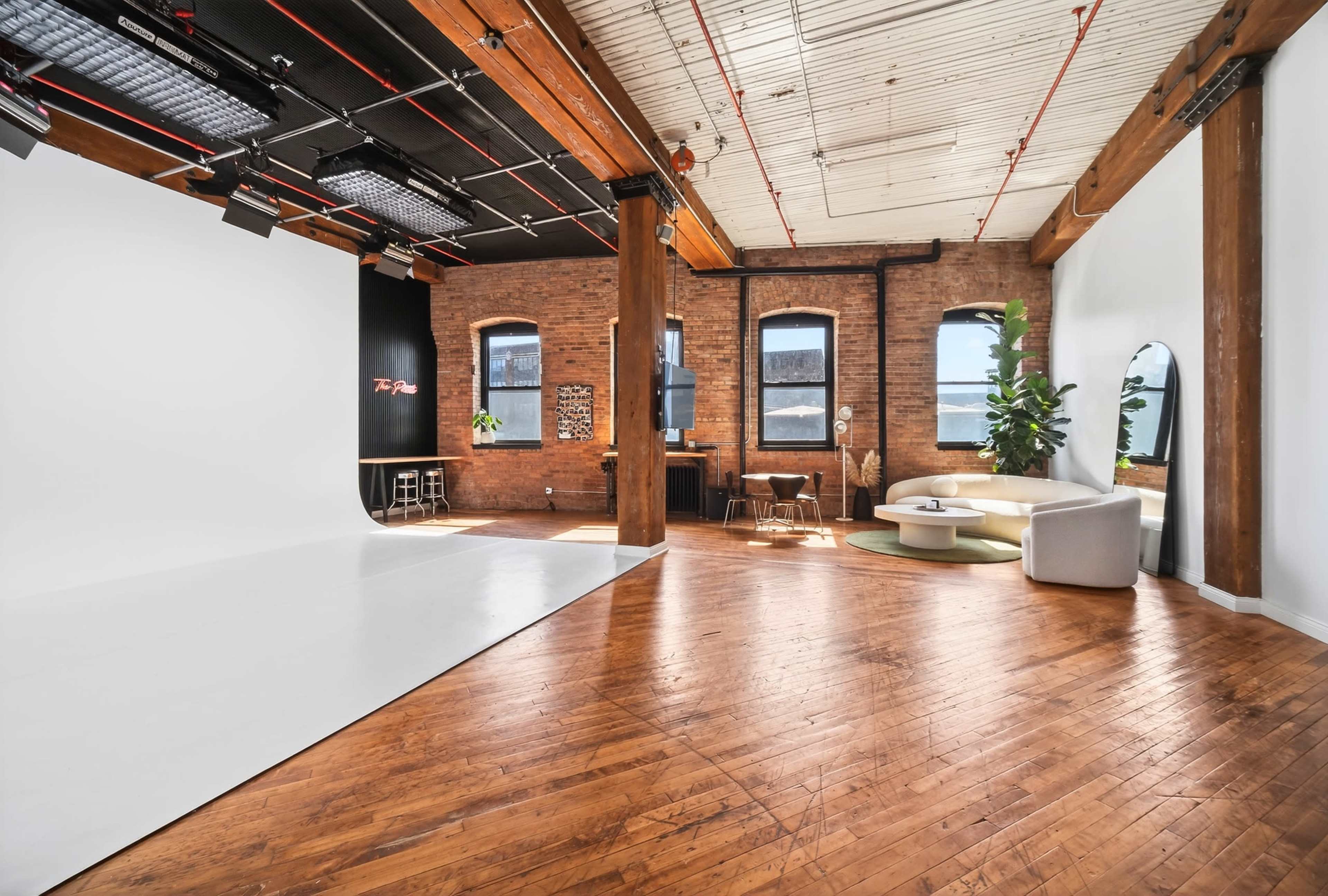 The image shows a spacious photo studio featuring wooden floors, exposed brick walls, and a large white backdrop, with a sectional couch and plants positioned in the corner.