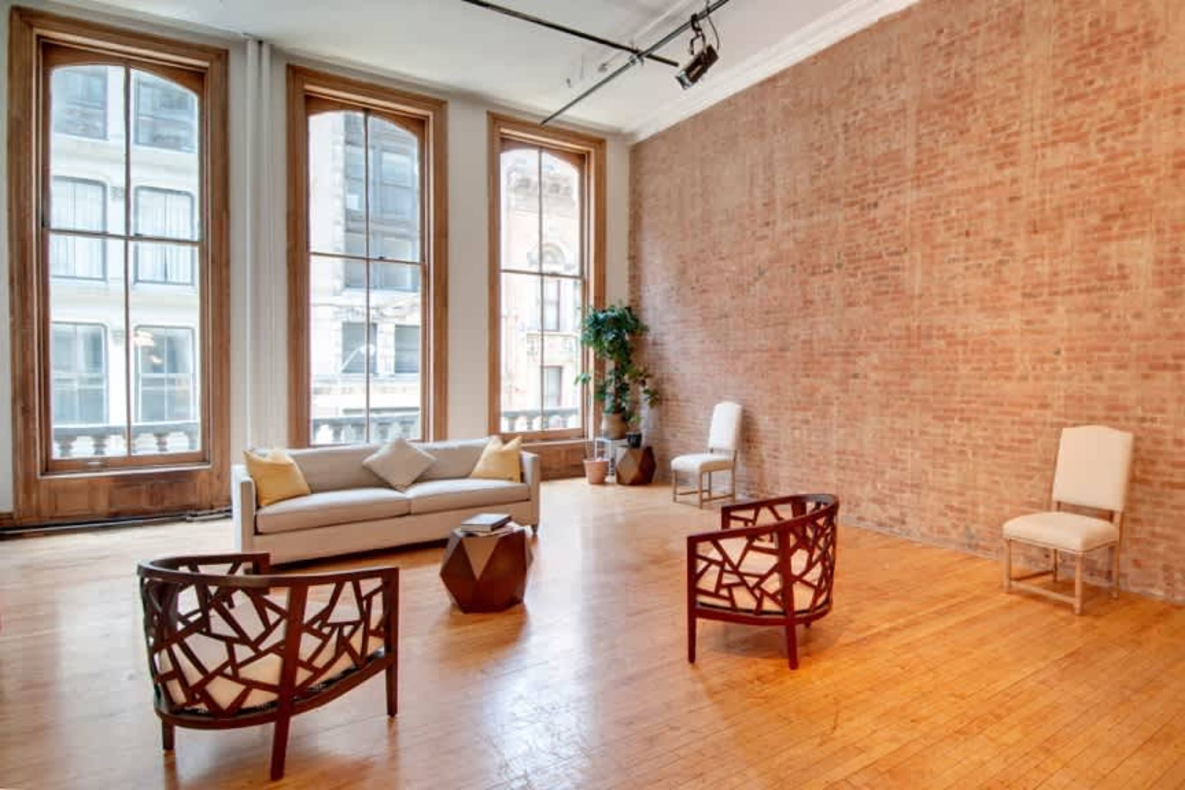The image shows a spacious room with large windows, featuring a brick wall, a sofa, two armchairs, and a small table in the center.