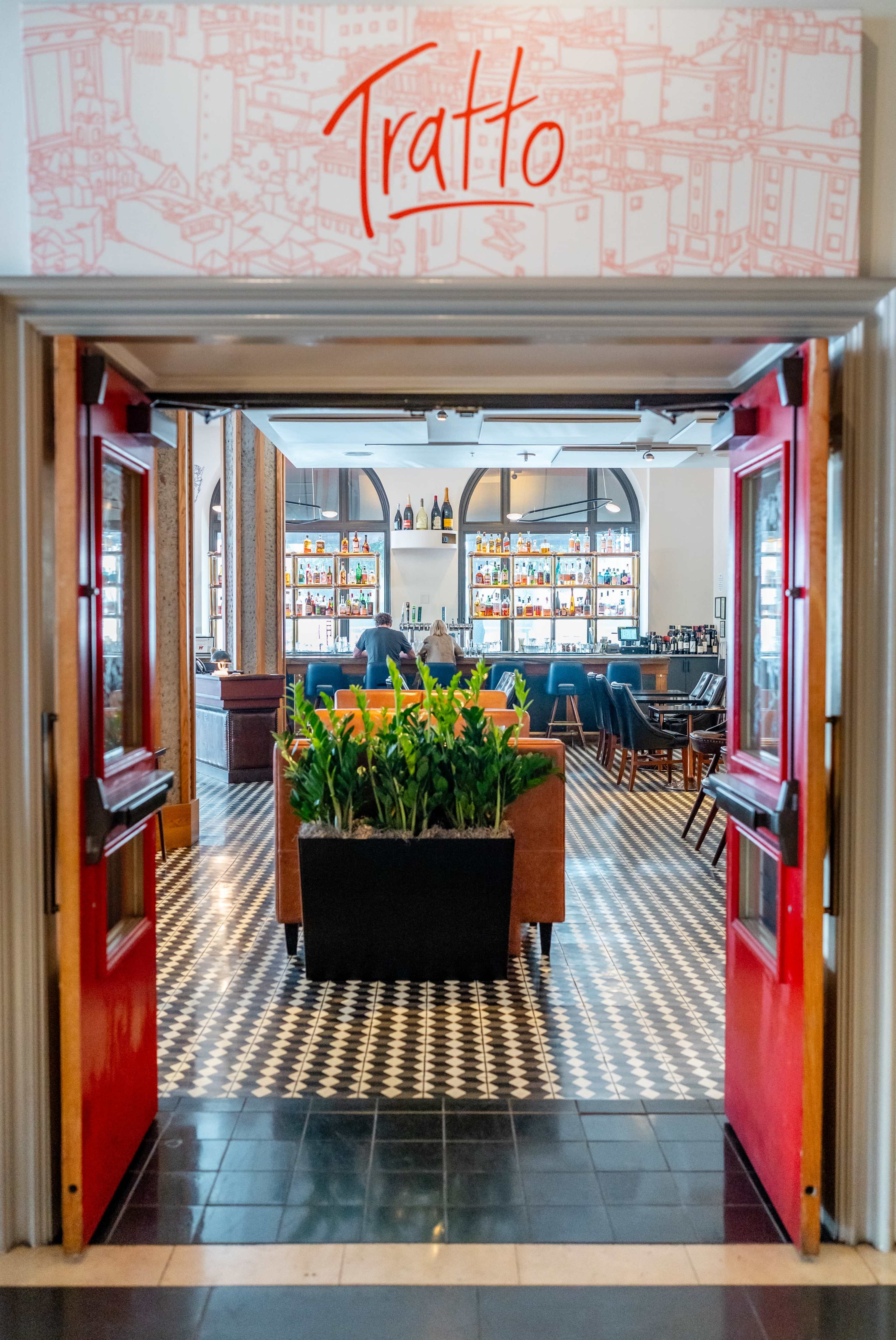 The image shows the entrance to a restaurant named "Tratto," featuring red doors that lead into a well-decorated dining area with a bar and seating.