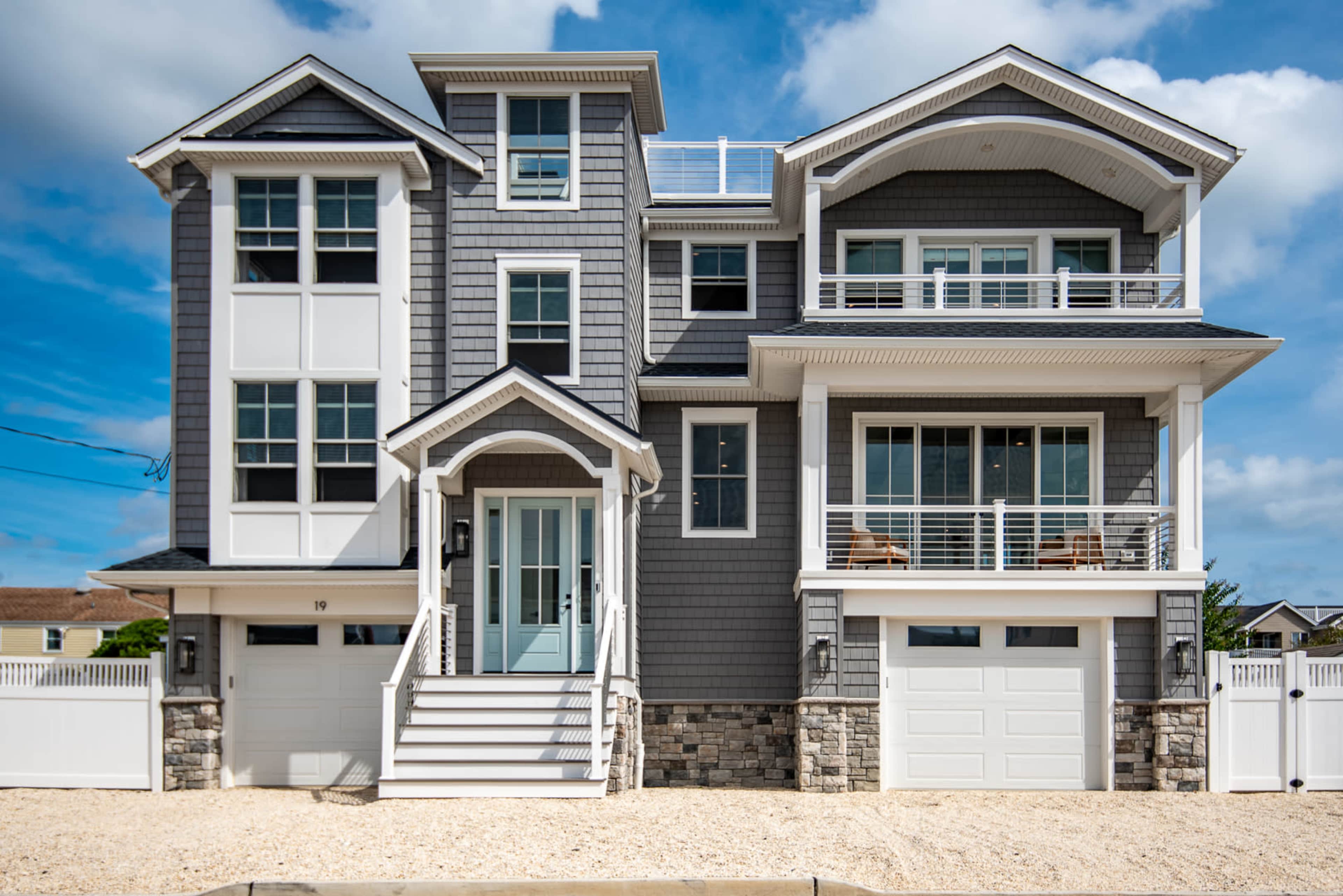A modern gray-shingled house featuring multiple levels, large windows, and two garage doors, surrounded by a sandy area and a white fence.