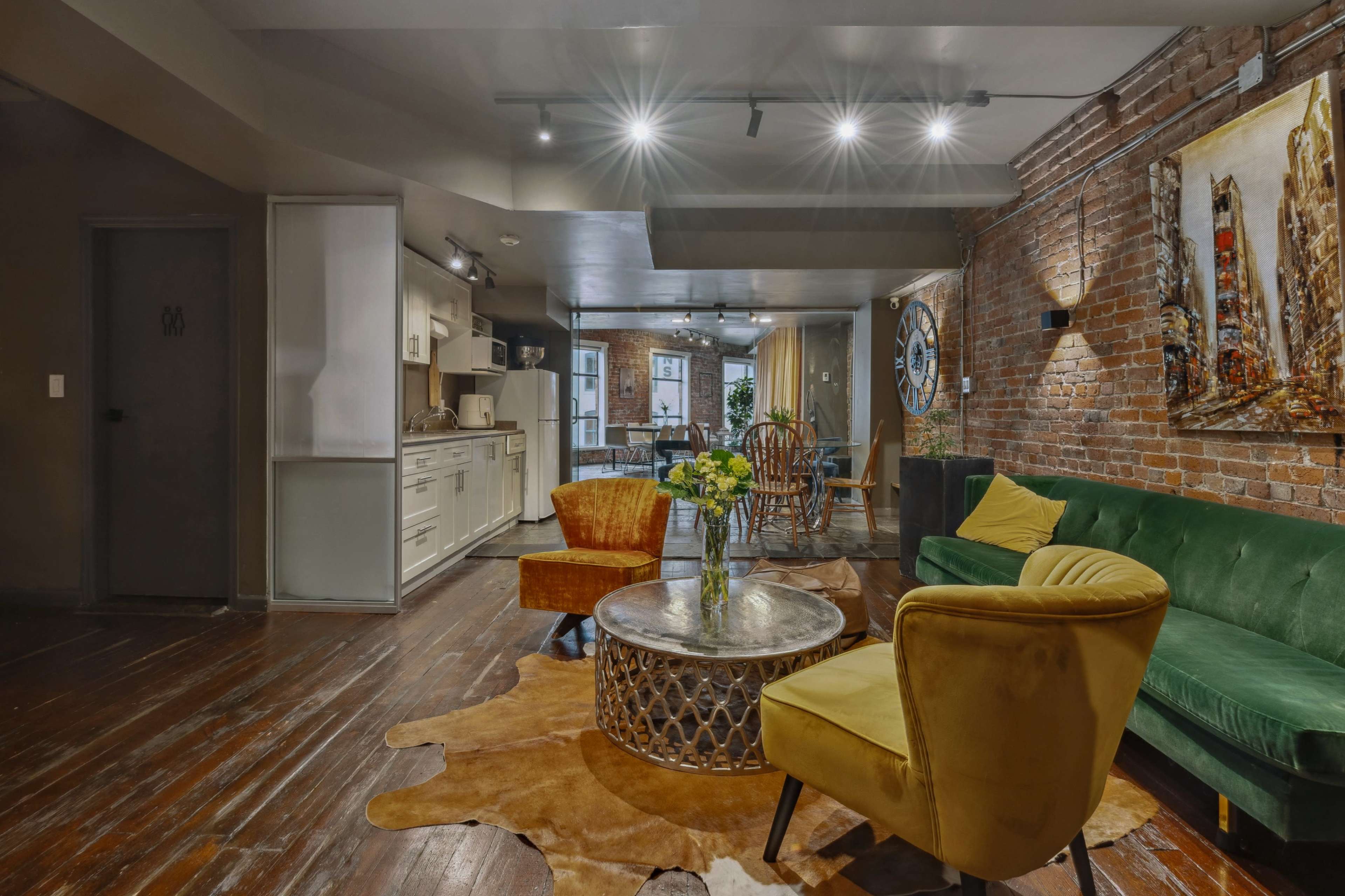 A modern living room features a green couch, yellow chairs, a round coffee table, and exposed brick walls, complemented by a decorative painting and plants.