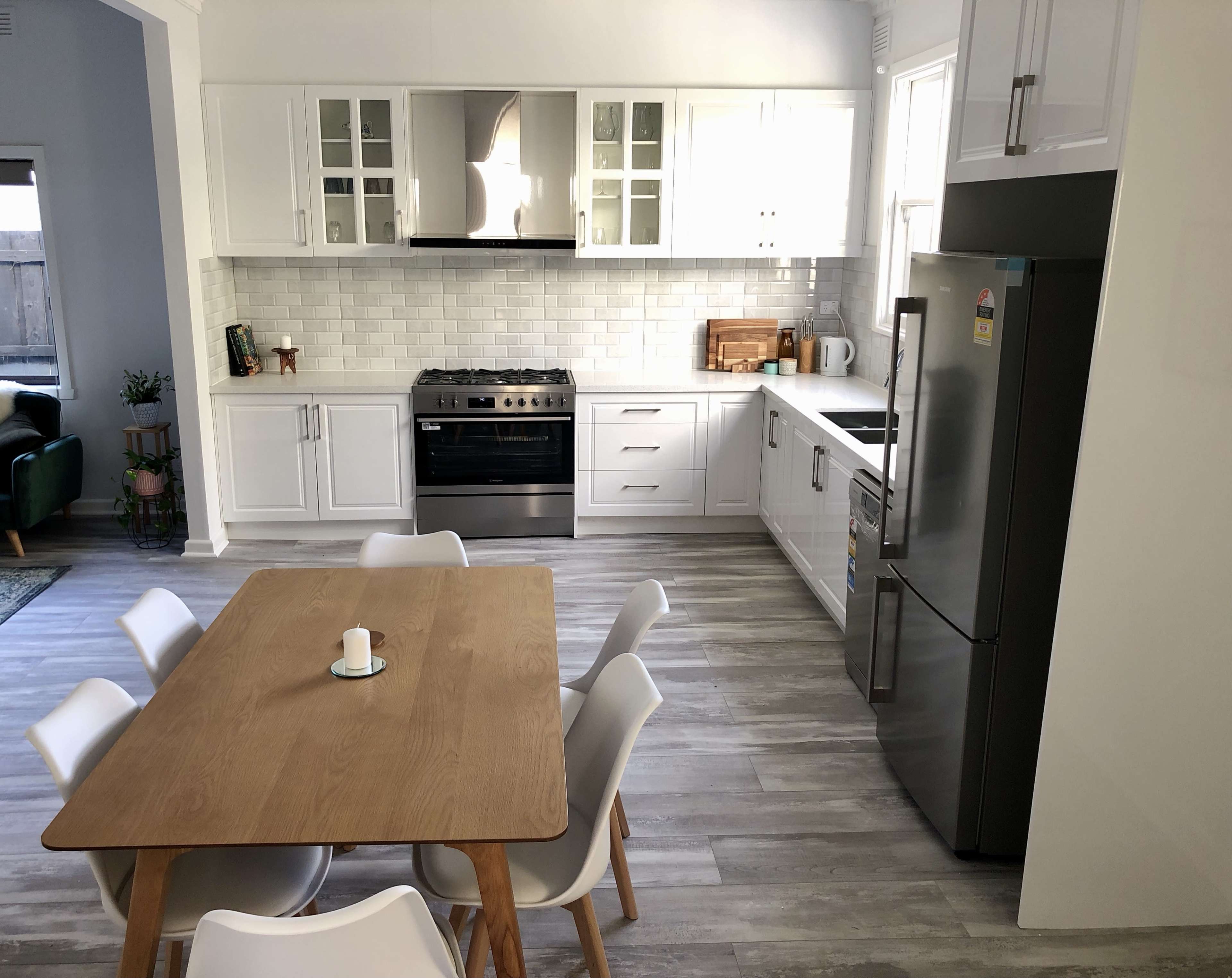 The image shows a modern kitchen with white cabinetry, a stainless steel refrigerator, a silver stove, and a wooden dining table surrounded by white chairs.