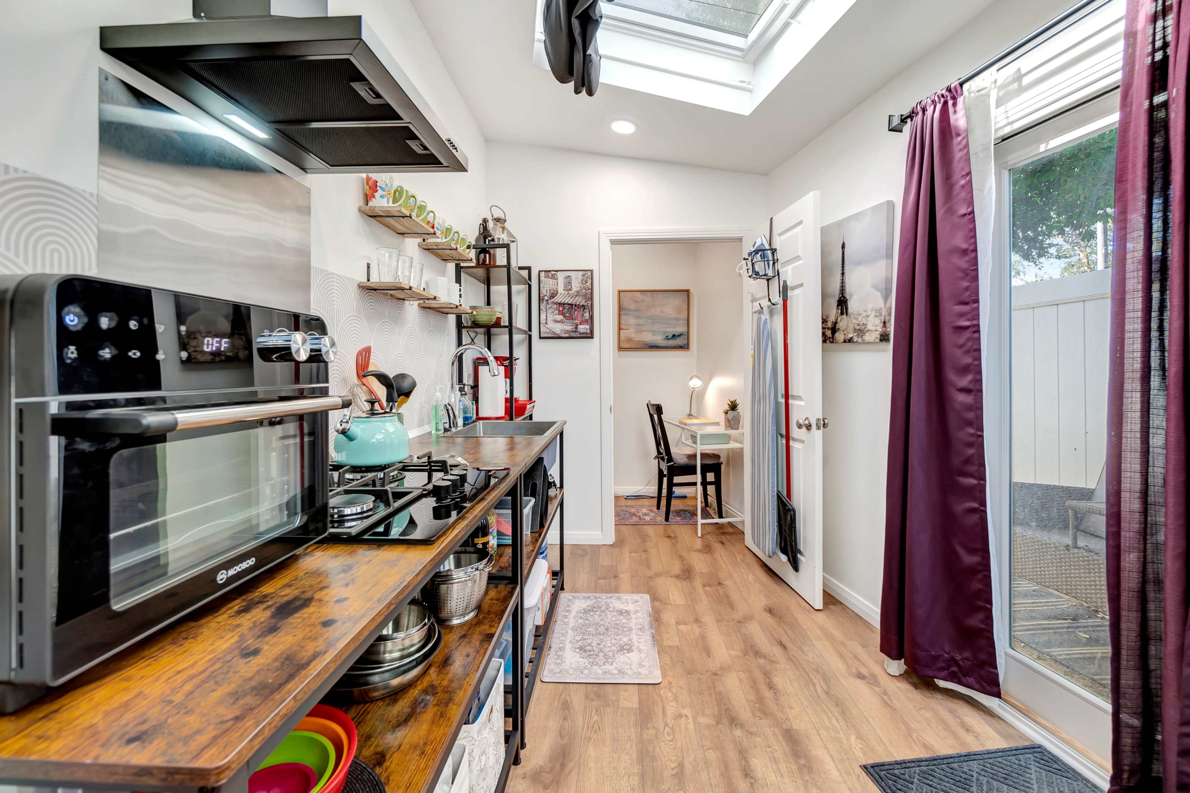 The image depicts a small kitchen area with modern appliances, wooden countertops, and a dining space visible through an open door.