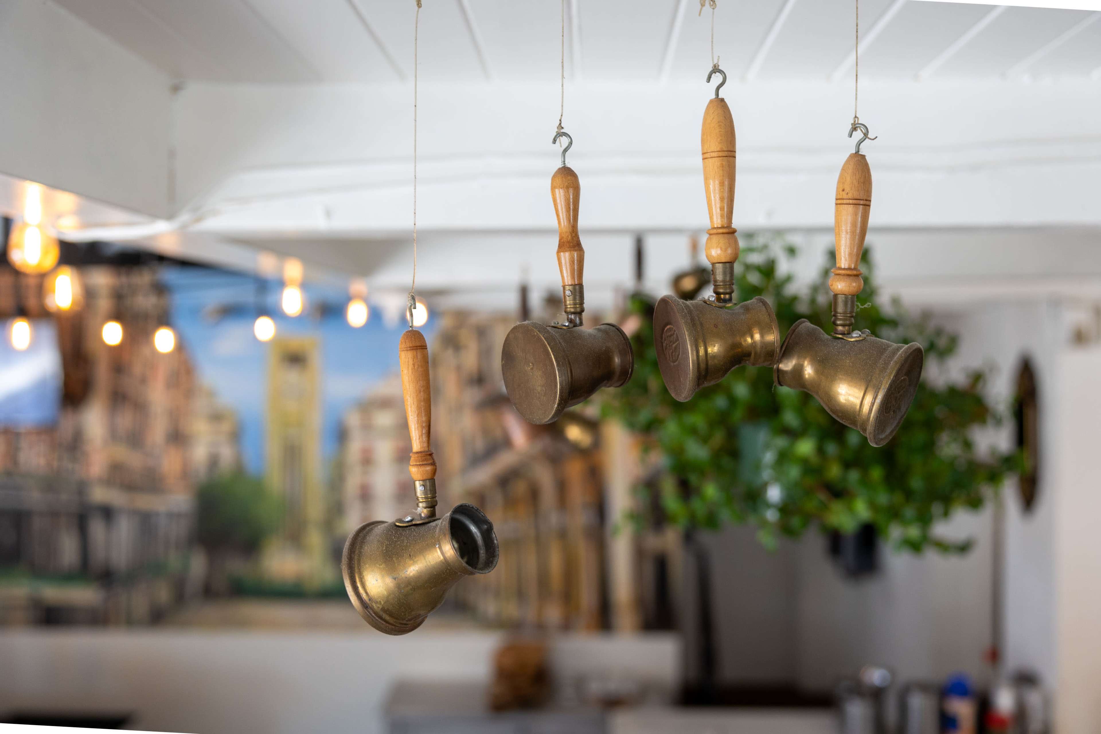 Brass coffee pots with wooden handles hang from a white ceiling in a café decorated with a city mural.