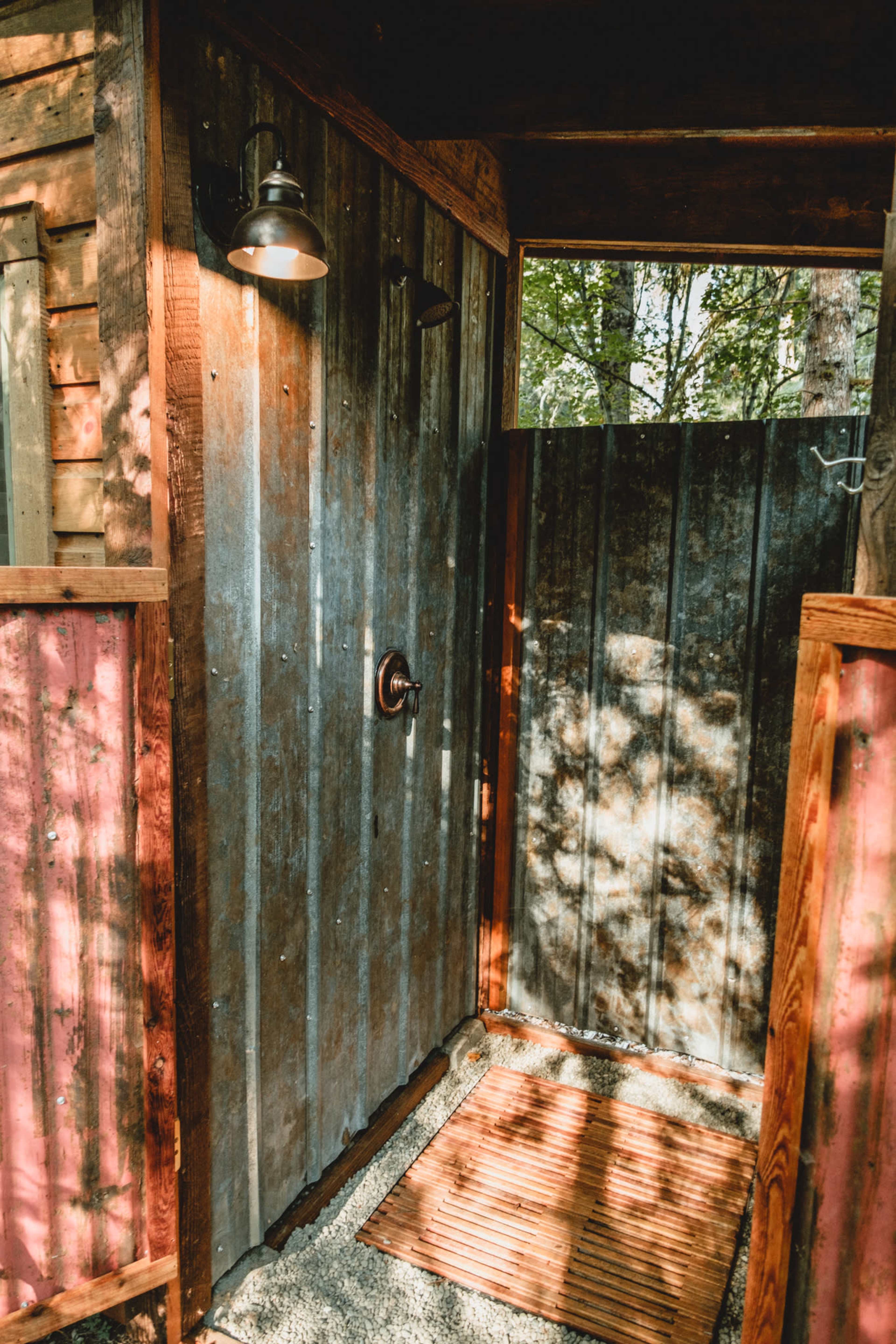An outdoor shower area enclosed by wooden walls, featuring a metal showerhead, a wooden mat on the ground, and a light fixture above.