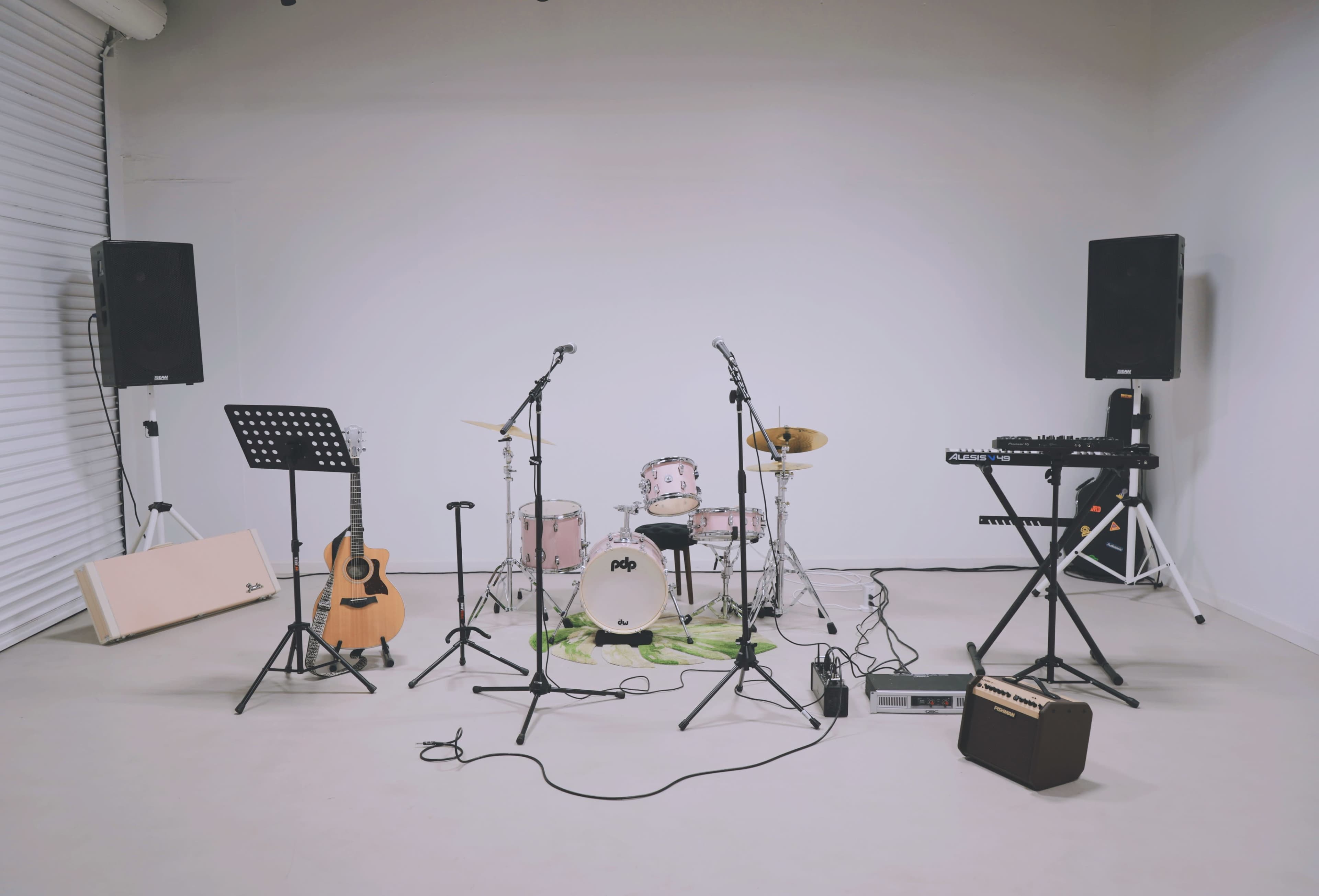 The image shows a music setup in a blank studio space, featuring a drum kit, guitars, and various microphones and musical equipment.