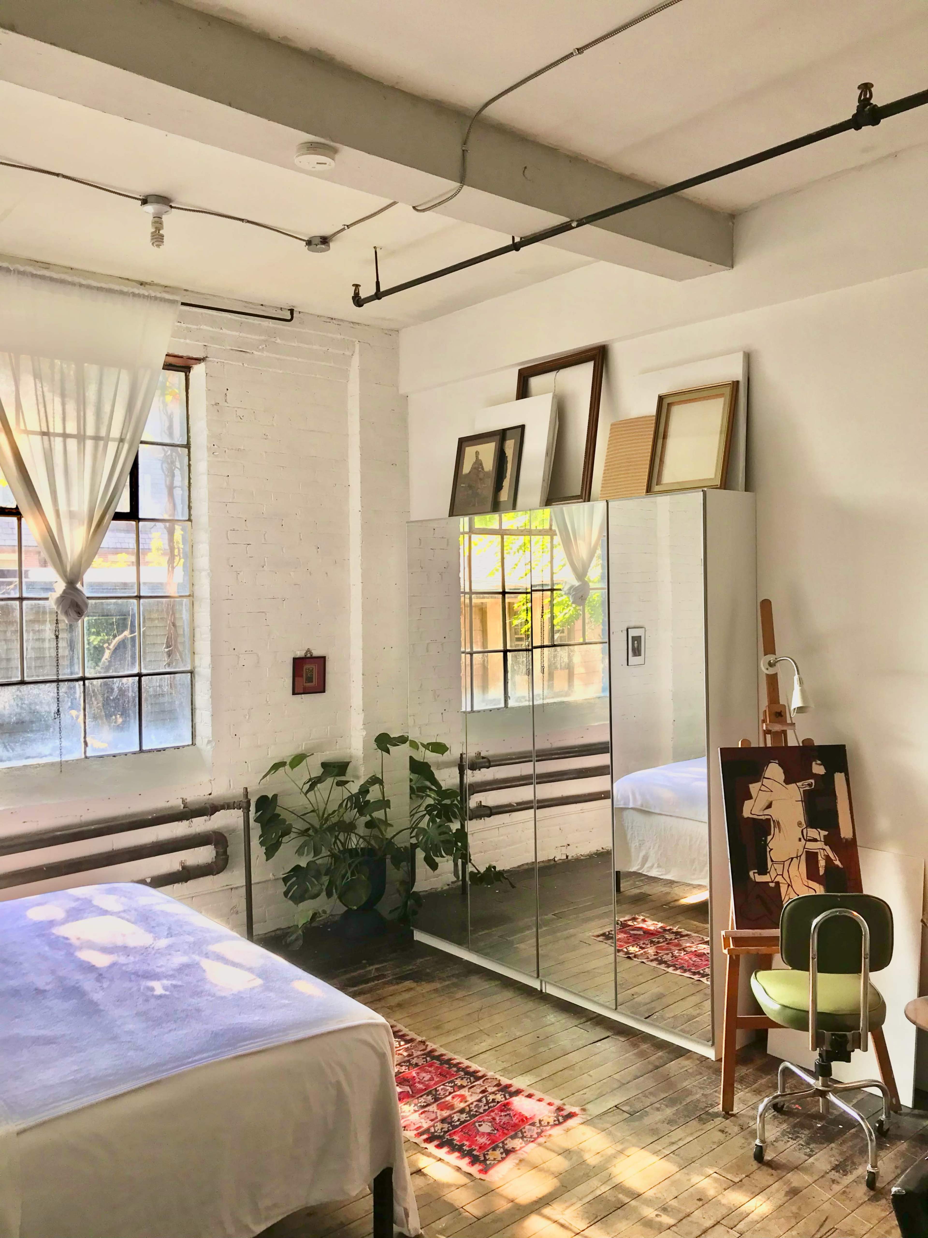 A bedroom featuring a bed, a mirror wardrobe, a small easel, and a potted plant, with light streaming in through large windows.