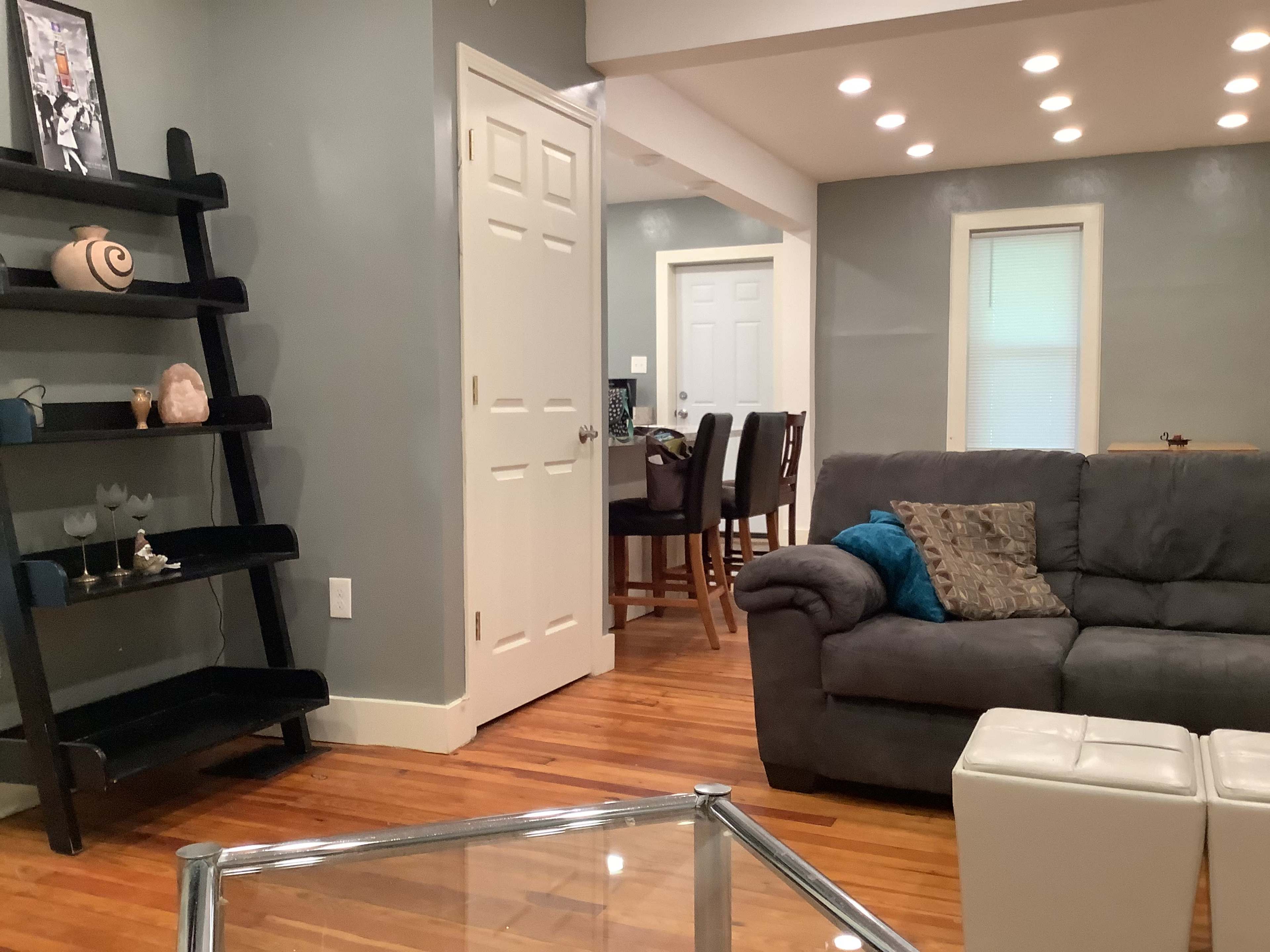 A cozy living room with a gray couch, a glass coffee table, and a black bookshelf against a light gray wall.