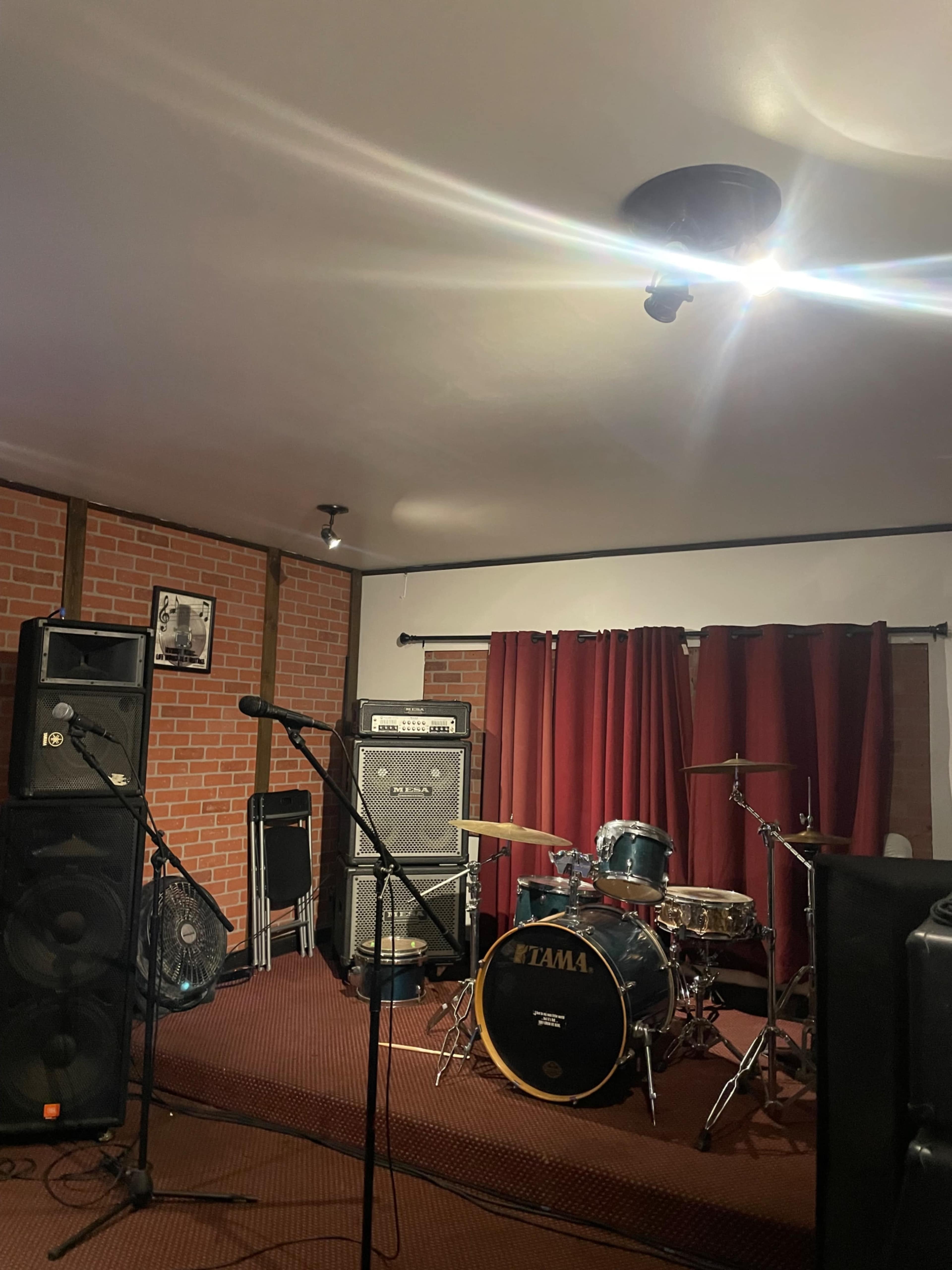The image shows a music rehearsal space featuring a drum set, amplifiers, microphones, and red curtains.