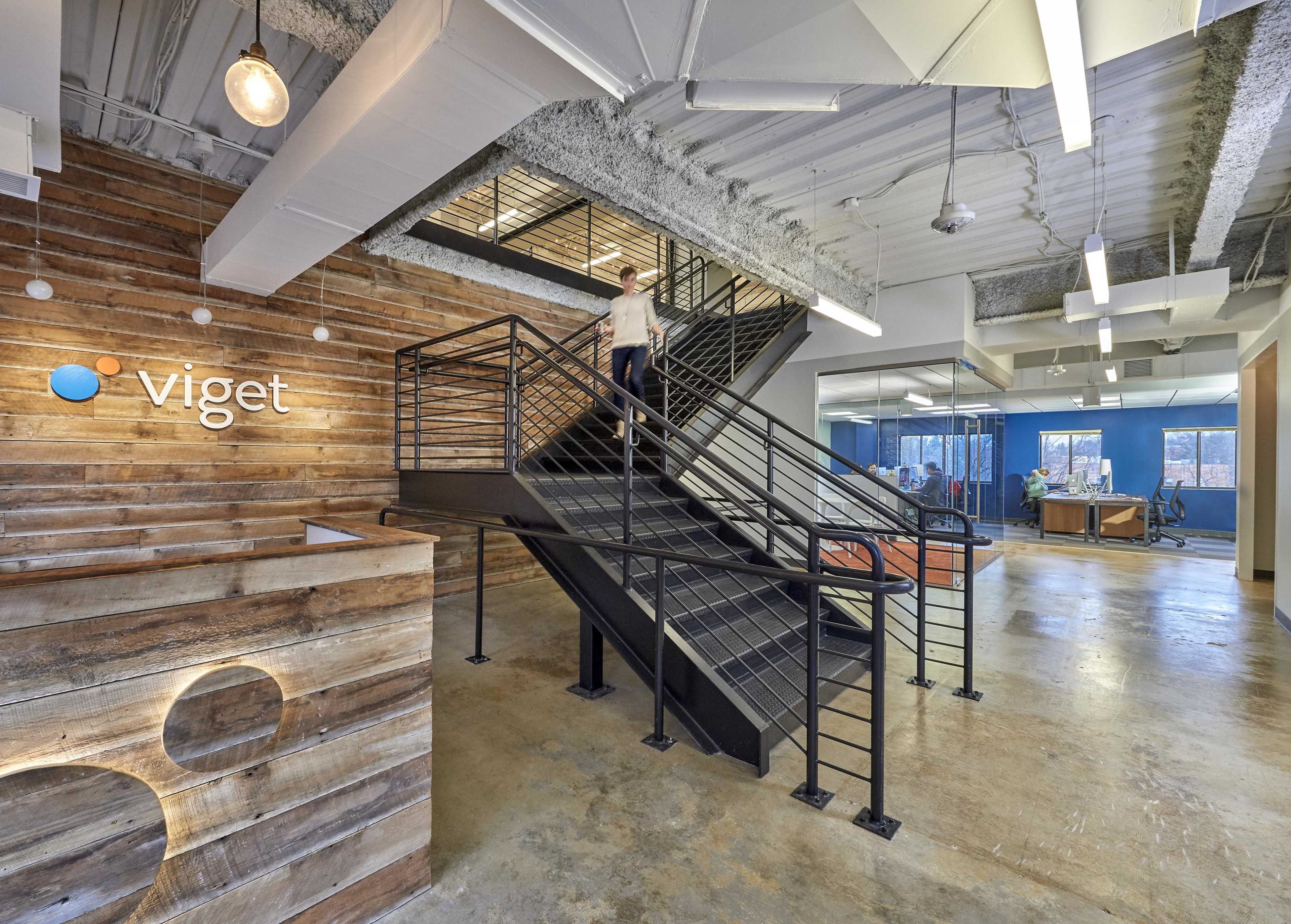A modern office lobby with a wooden reception desk and a staircase leading to an upper level, featuring exposed ceiling beams and contemporary lighting.